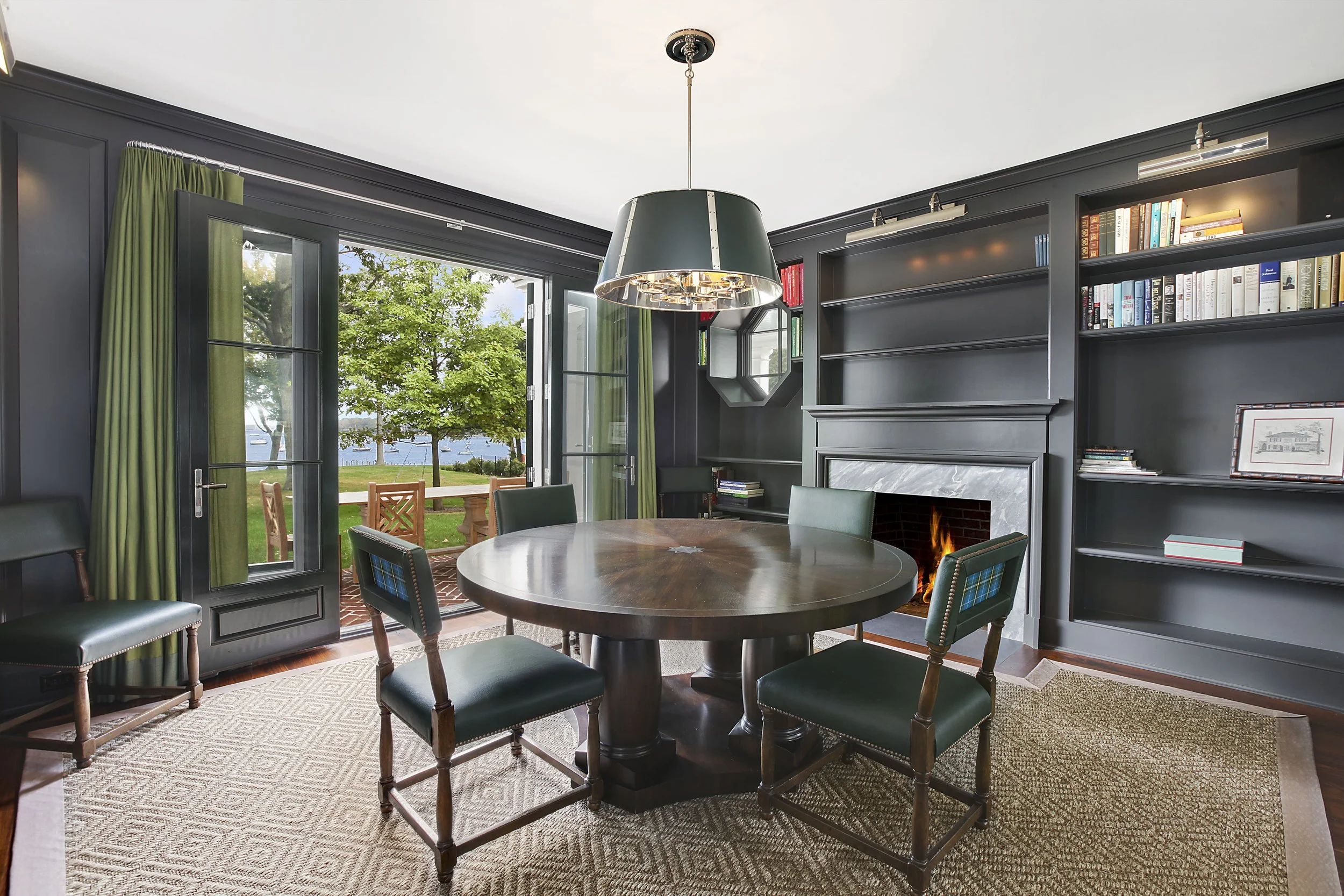 Interior view of a dining room with a round wooden table, four green upholstered chairs, a fireplace with a gray mantel, built-in bookshelves, green curtains, and an open door revealing a view of a lake and trees outside.