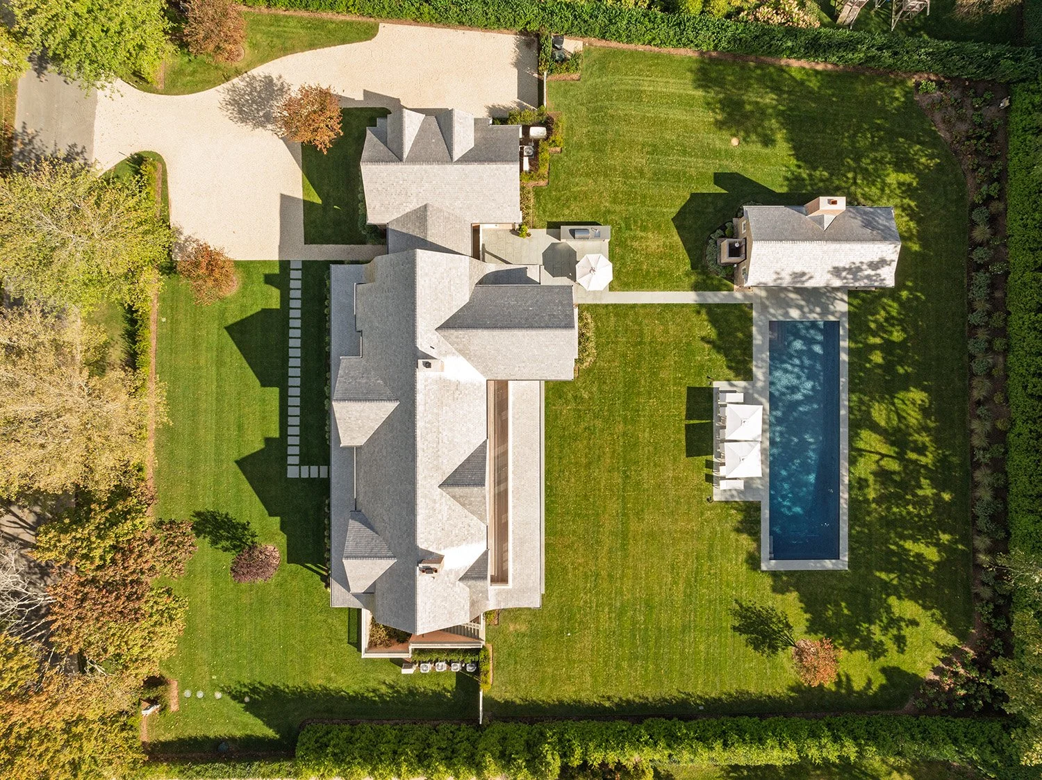 Aerial view of a large residential backyard featuring a house with multiple roofs, a rectangular swimming pool, a lawn with trees, a small shed or guesthouse, and a paved driveway lined with trees. Construction in Hamptons New York