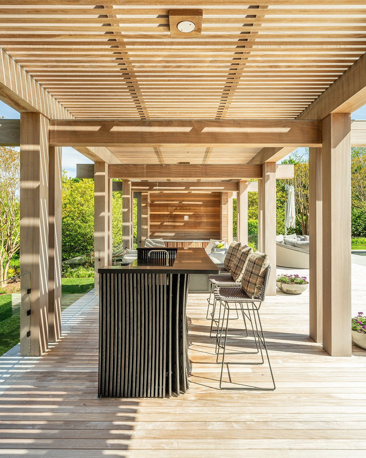 Outdoor wooden patio with a bar counter, plaid cushioned chairs, lounge seating, and potted plants, surrounded by greenery.