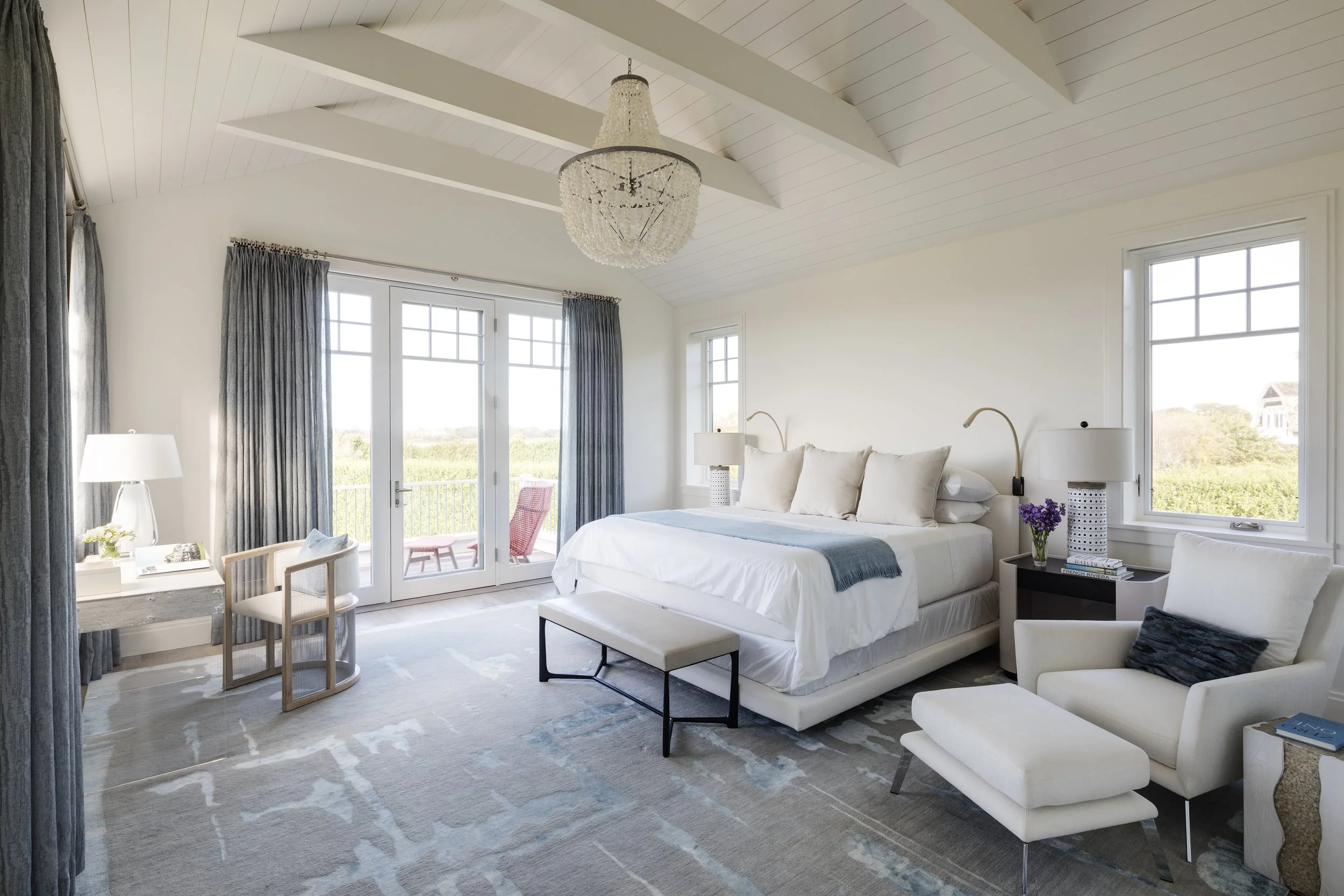 Bright, spacious bedroom with a large bed, white bedding, and multiple pillows. There are large windows and glass doors leading to a balcony with a pink chair. The room has a chandelier, a chair and a desk with a lamp and decorative items, and a sitt