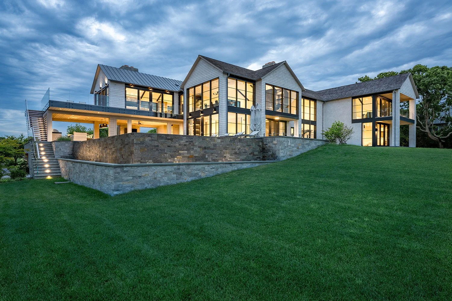 Modern two-story house with large glass windows, surrounded by a well-manicured lawn and an outdoor patio at dusk.