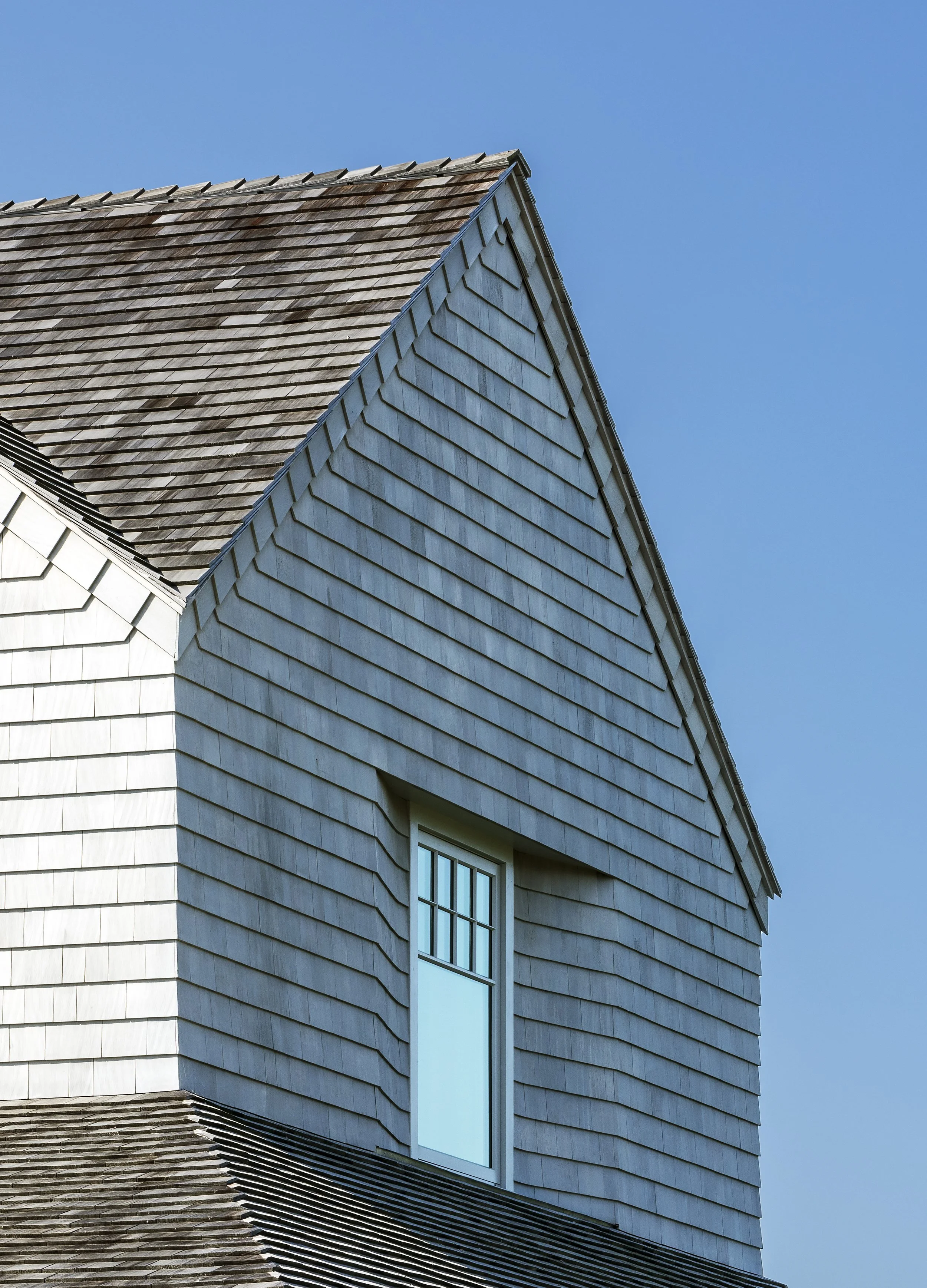 Close-up of a house with gray wooden shingles, a brown tiled roof, and a single window against a clear blue sky.