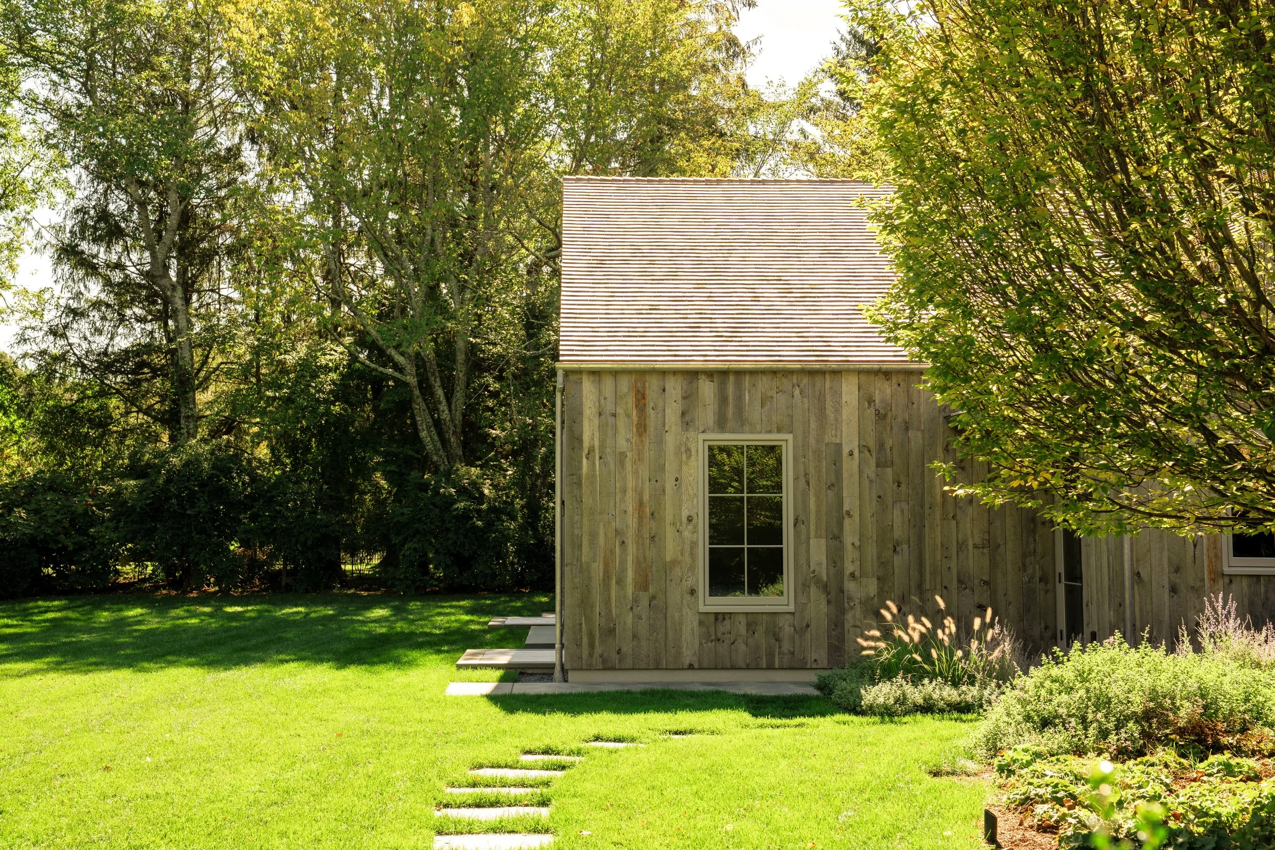 A small wooden house with a gabled roof surrounded by a green lawn, trees, and bushes.