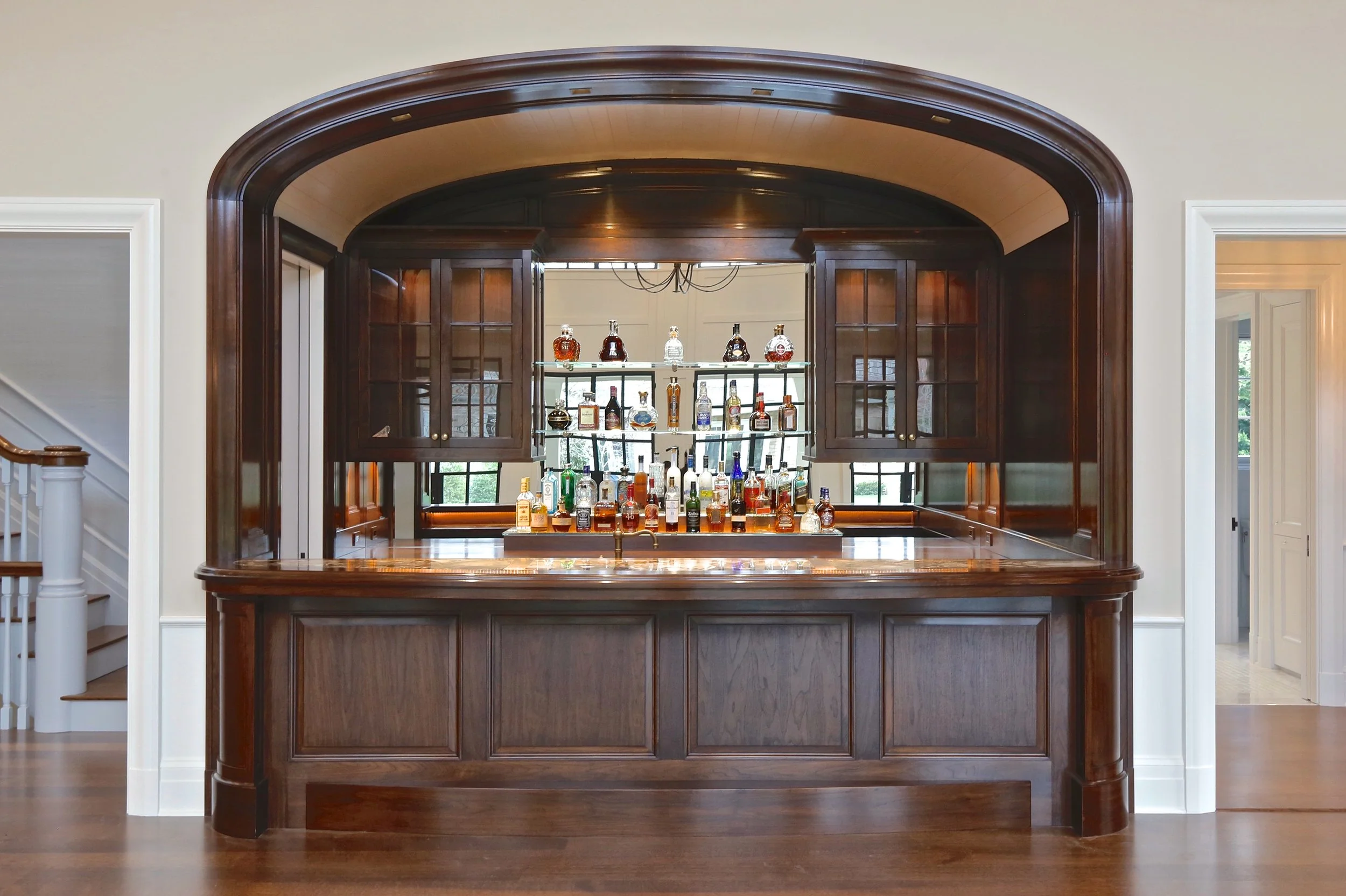 A home bar with a dark wooden finish, situated behind an arched opening in a room with white walls, hardwood flooring, and a doorway on each side. The bar has bottles of alcohol on shelves and a countertop.