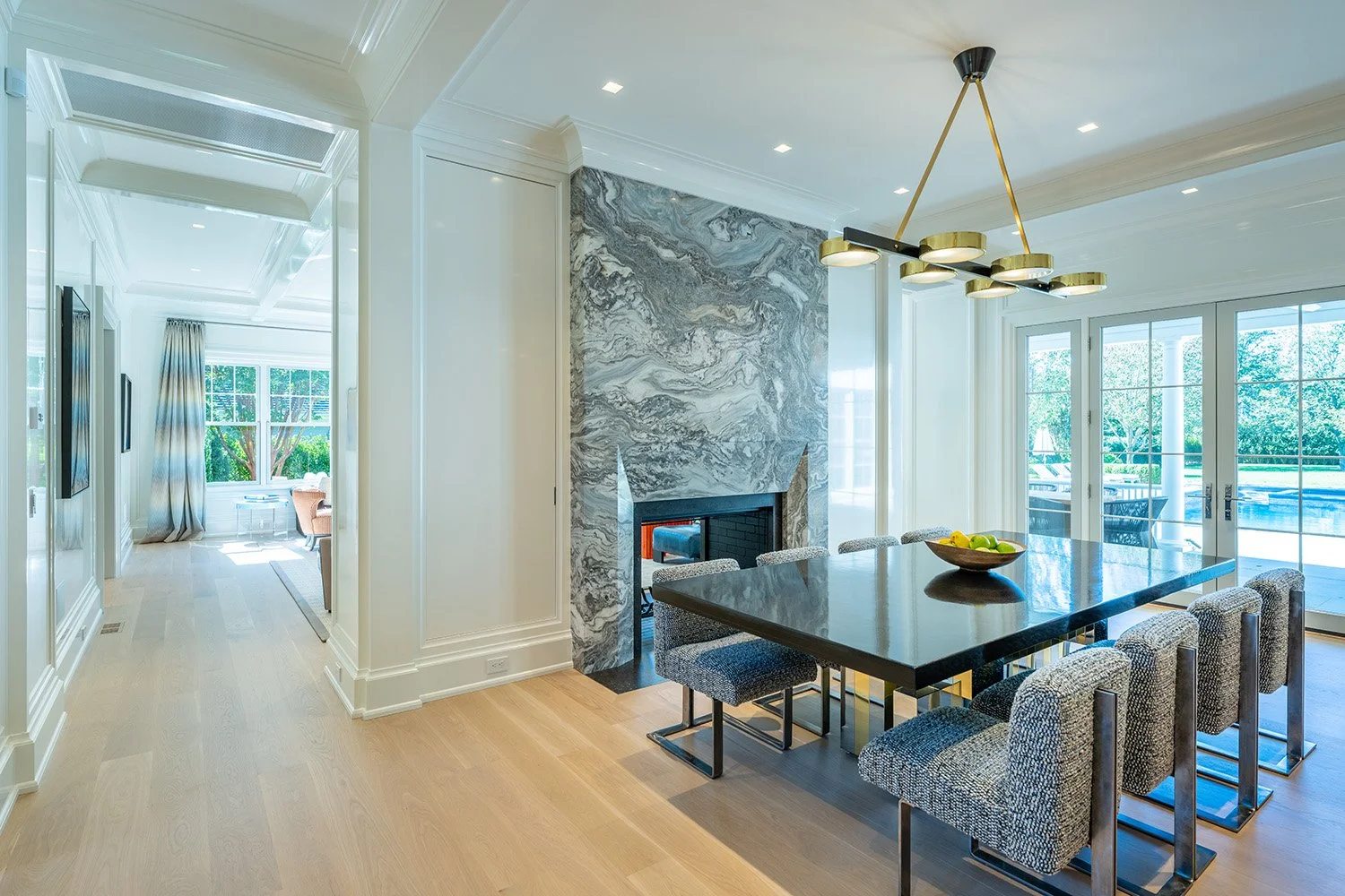 Modern dining room with a black marble table, eight upholstered chairs, a marble fireplace, and large glass sliding doors leading to an outdoor patio with greenery.