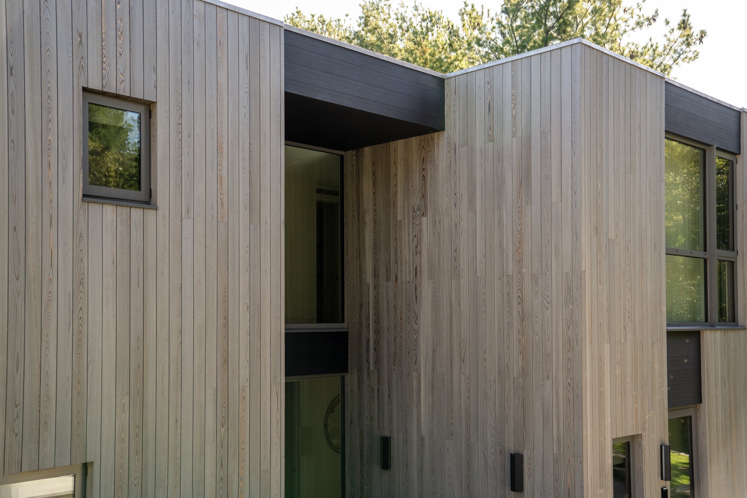 Close-up of a modern building with vertical wooden siding and black window frames, with trees in the background.