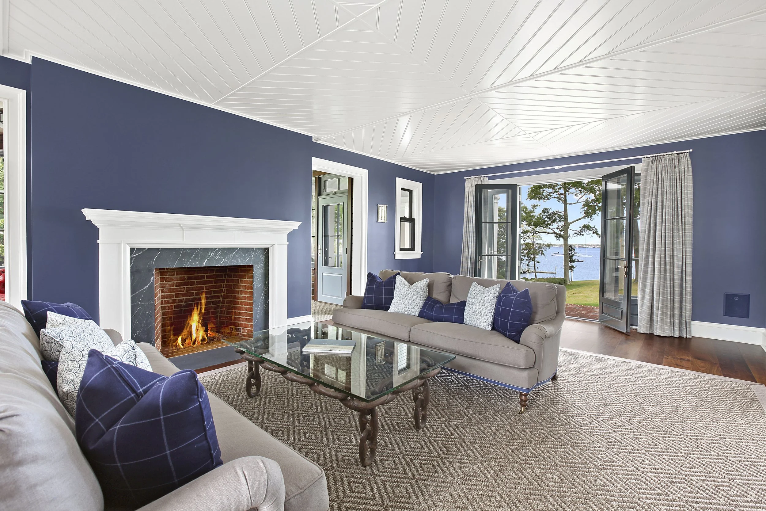 Living room with blue walls, white ceiling, beige sofas with blue and white throw pillows, glass coffee table, fireplace with fire, window views of water and trees, open doors with curtains.