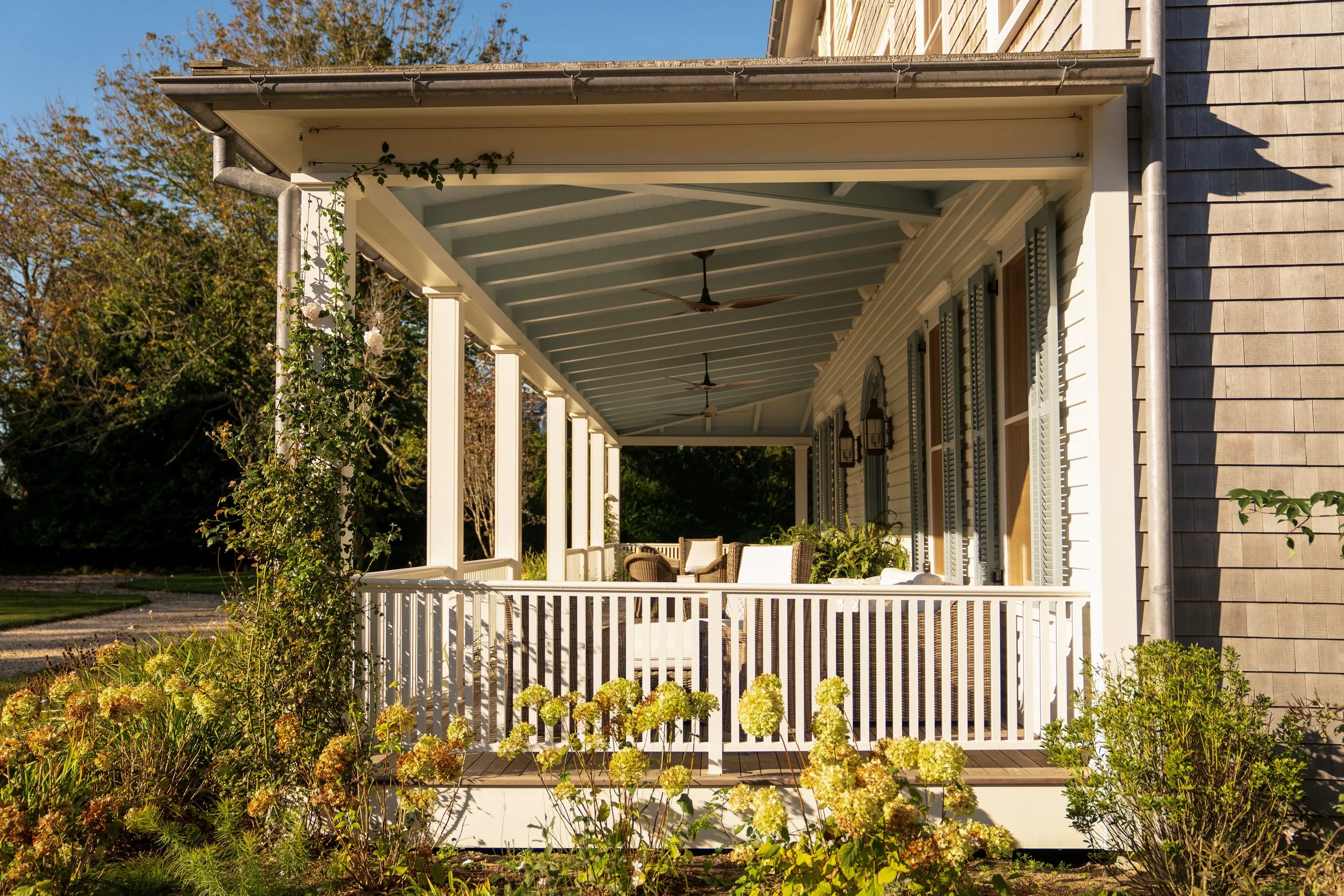 Front porch of a house with white railing, outdoor furniture, and ceiling fans, surrounded by plants and trees.