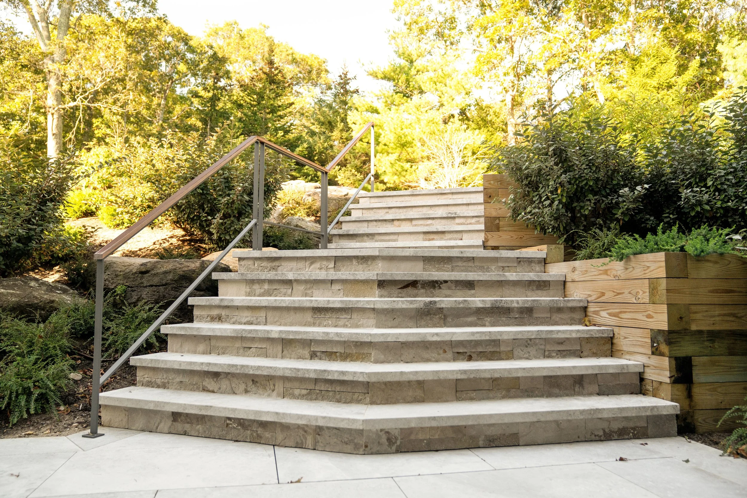 Outdoor stone staircase with metal handrail leading up a landscaped garden with bushes and trees.