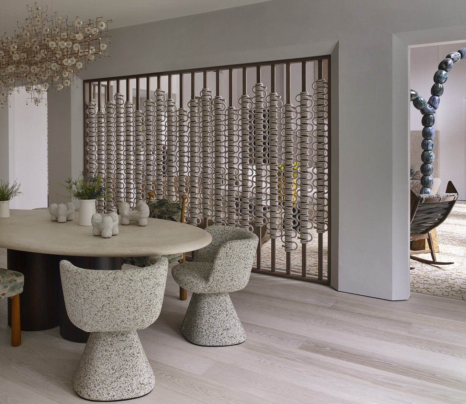 A dining area with a round table, four textured chairs, decorative vases with greenery, and a unique partition made of stacked rings, with a glimpse of an additional seating area with a column of large spherical objects.