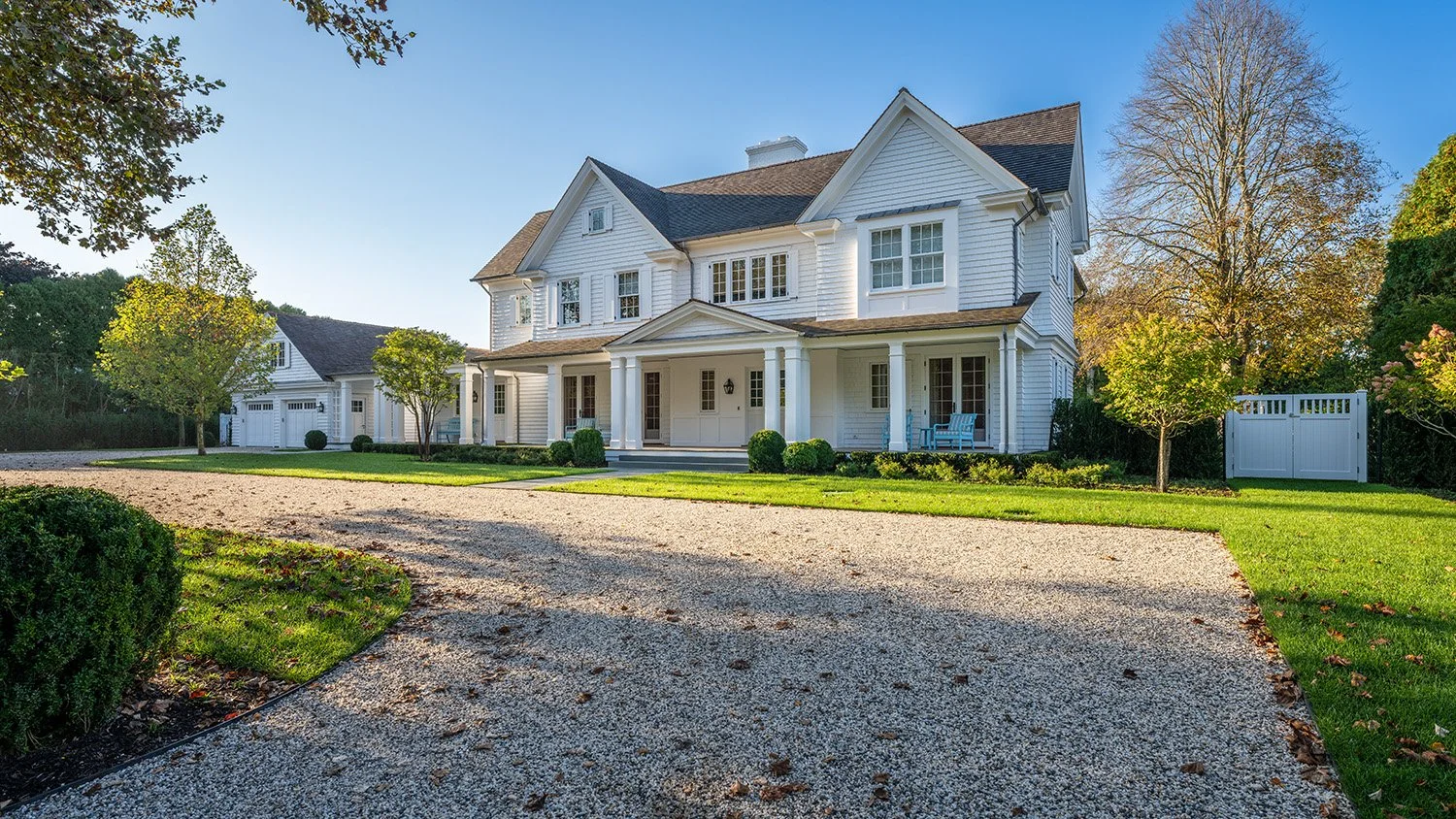 A large white house with a brown roof, multiple windows, and a front porch with benches. There is a gravel driveway leading up to the house, surrounded by well-maintained green grass and trees, with a clear blue sky overhead.