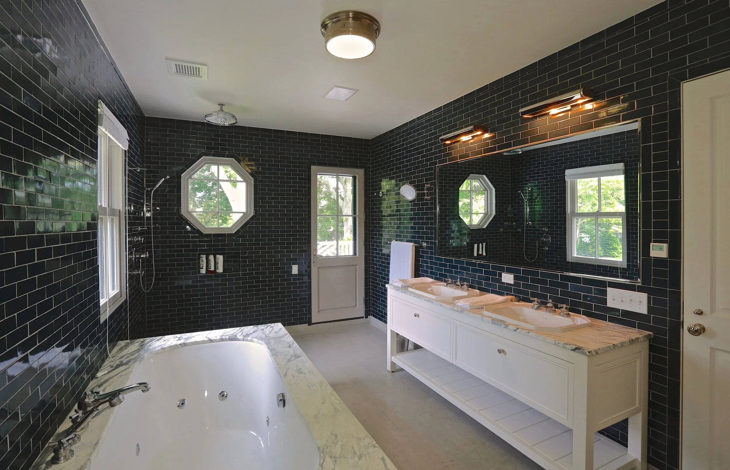 Bathroom with black tile walls, a large bathtub with a marble surround, a white double vanity with a marble countertop and two sinks, a large mirror, and two windows with greenery outside. There is a door leading outside and various fixtures like a s