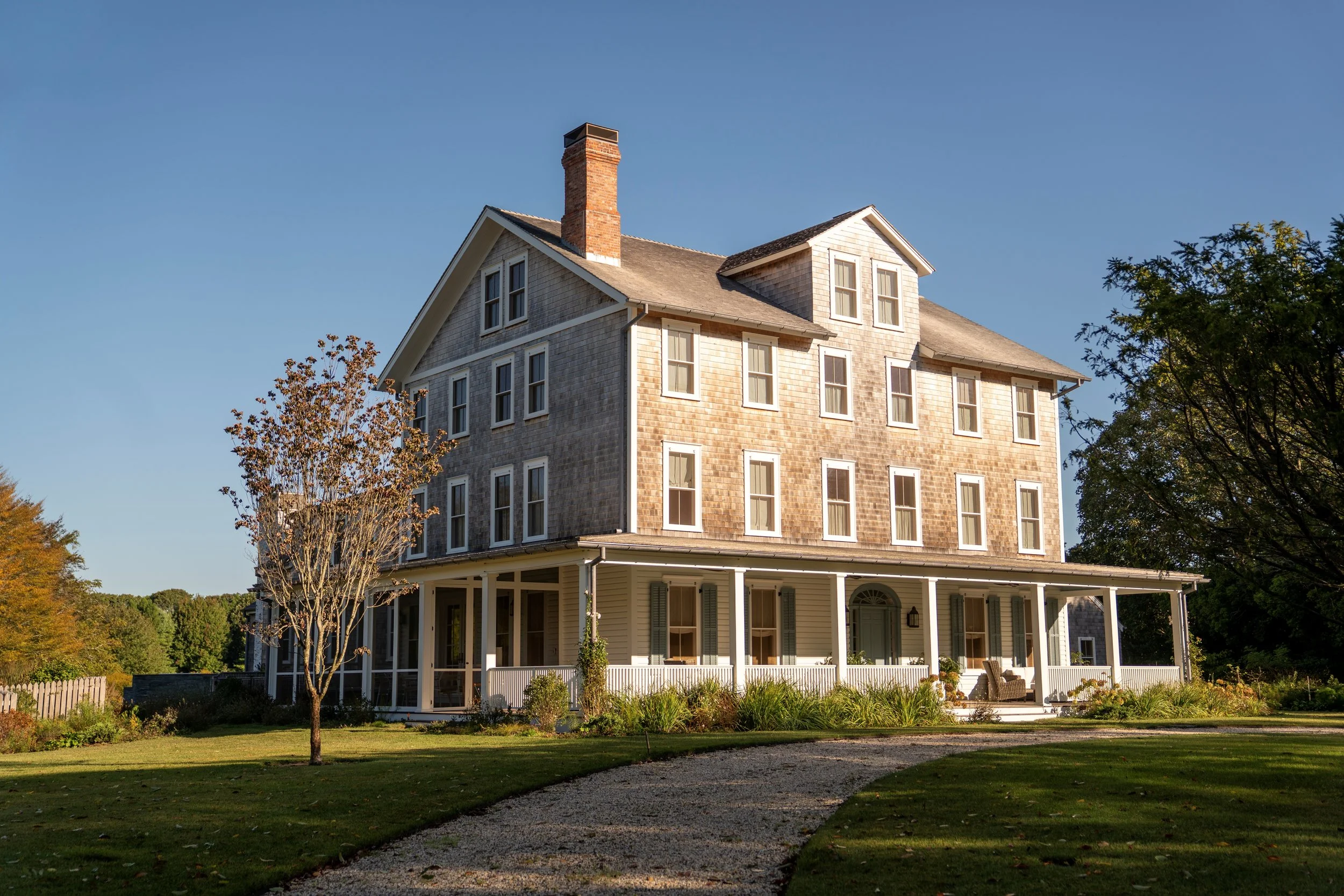 A large, multi-story house with a wraparound porch, situated on a grassy area with trees and a gravel pathway leading to the entrance, under a clear blue sky.
