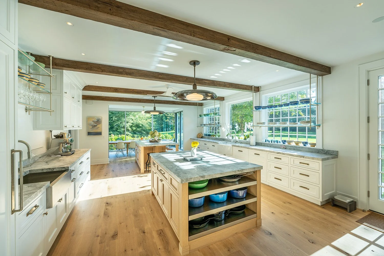 Bright kitchen with white cabinets, marble countertops, a wooden island with open shelves, and large windows showing a green outdoor view.