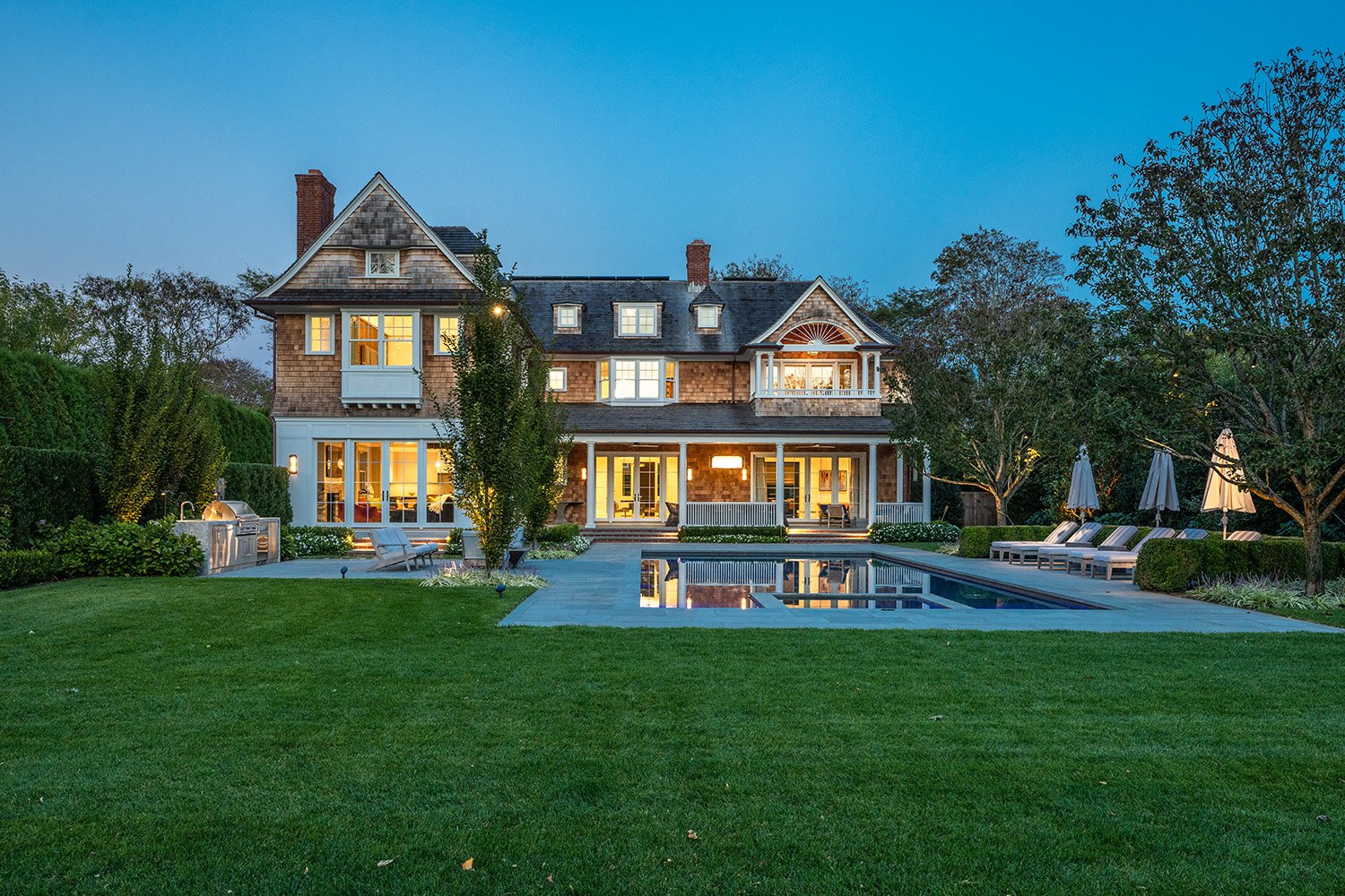 Large house with a backyard, swimming pool, lounge chairs, trees, and outdoor BBQ grill, illuminated at dusk. Custom residential construction in the Hamptons