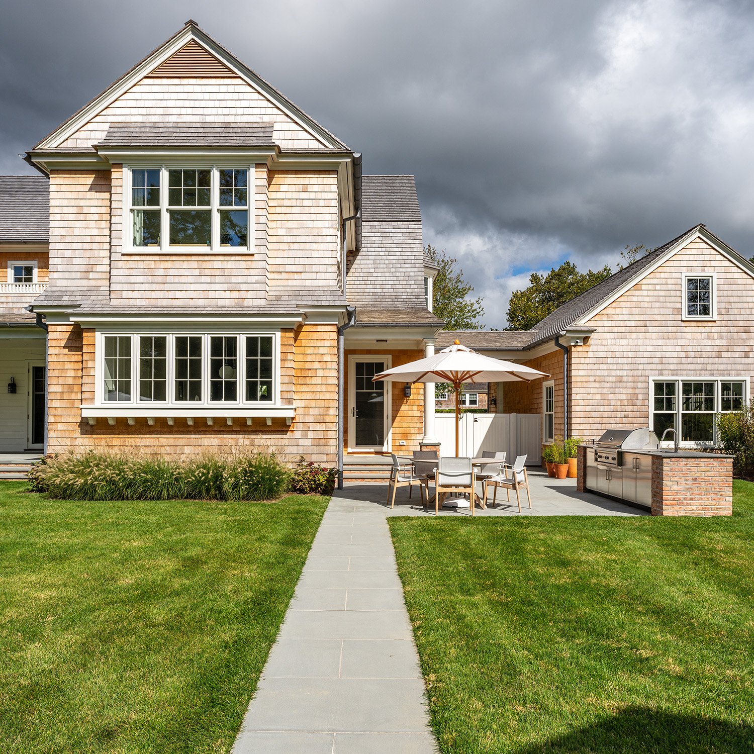 Backyard of a house with a patio set and grill, American style home with shingled exterior, green lawn, cloudy sky. Construction in Hamptons New York
