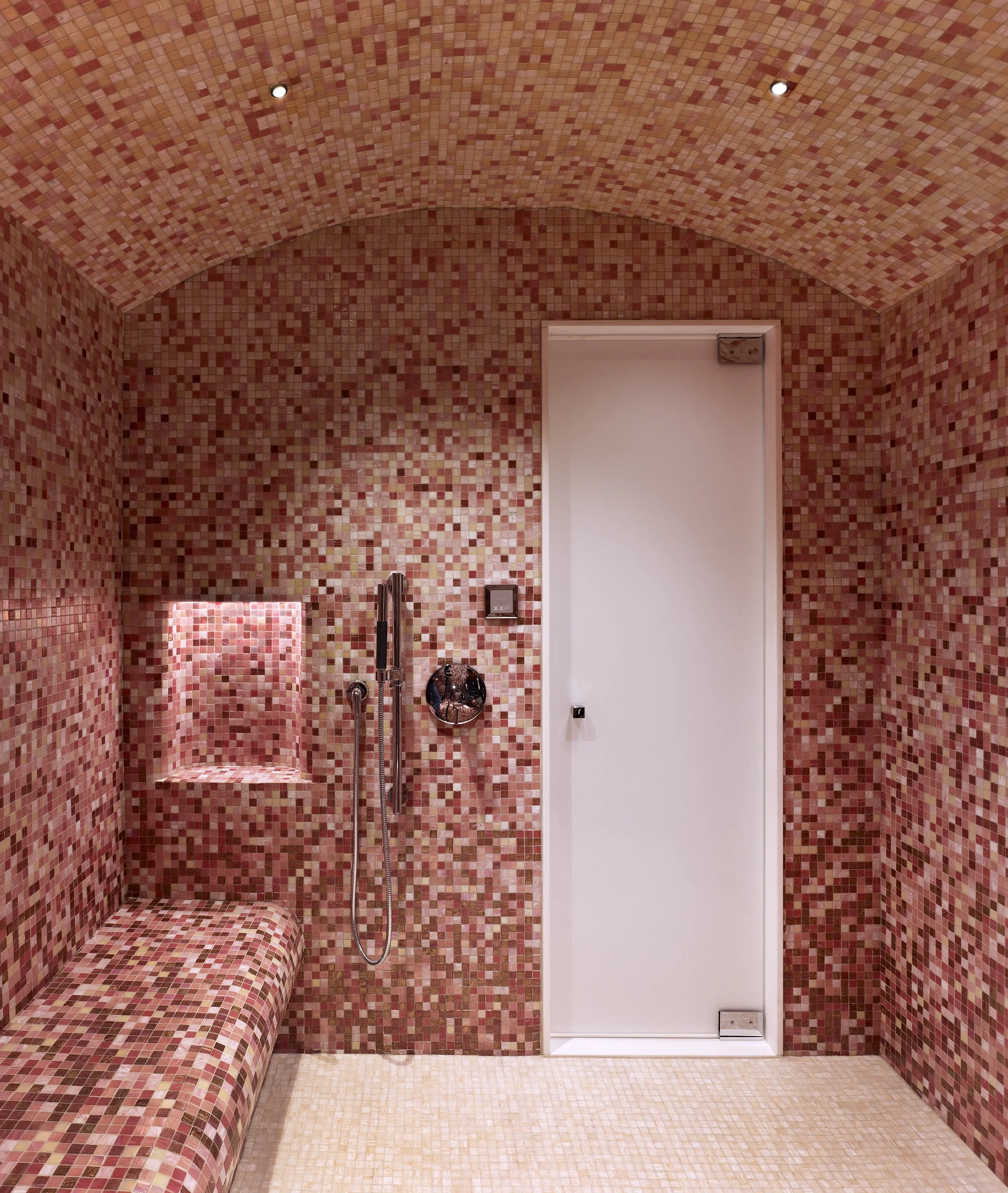 A mosaic tiled shower with a built-in bench, a niche with pink tiles, rain shower head, handheld shower, and a white door.