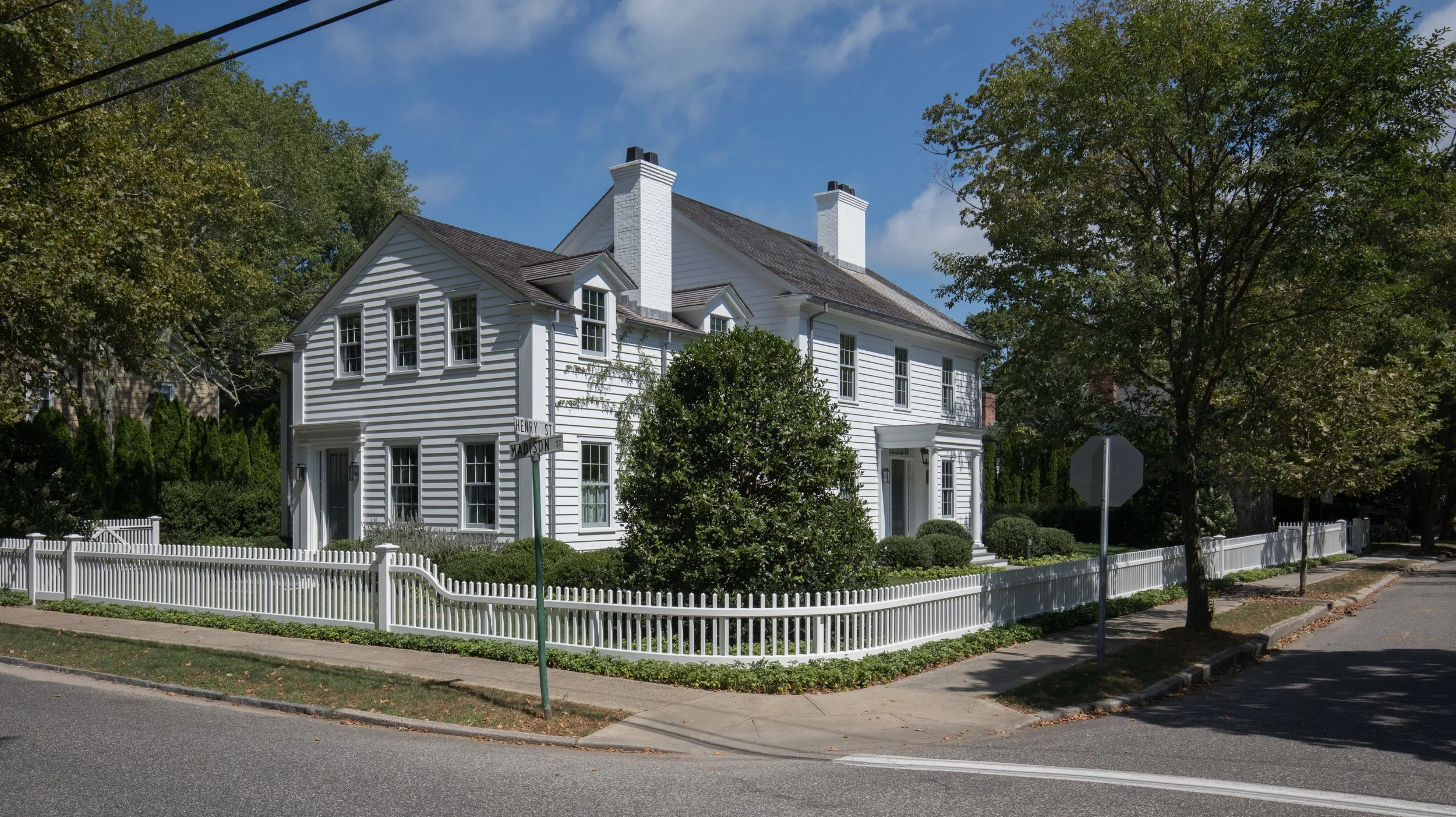 A white two-story house with a wrap-around porch, surrounded by a white picket fence and greenery, on a street corner under a partly cloudy sky. Sag Harbor