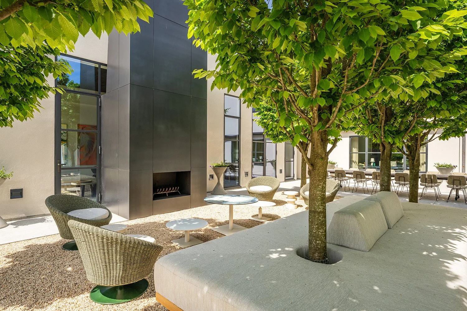 An outdoor patio area with trees, modern chairs, a fireplace, and a seating area near a building with large windows.
