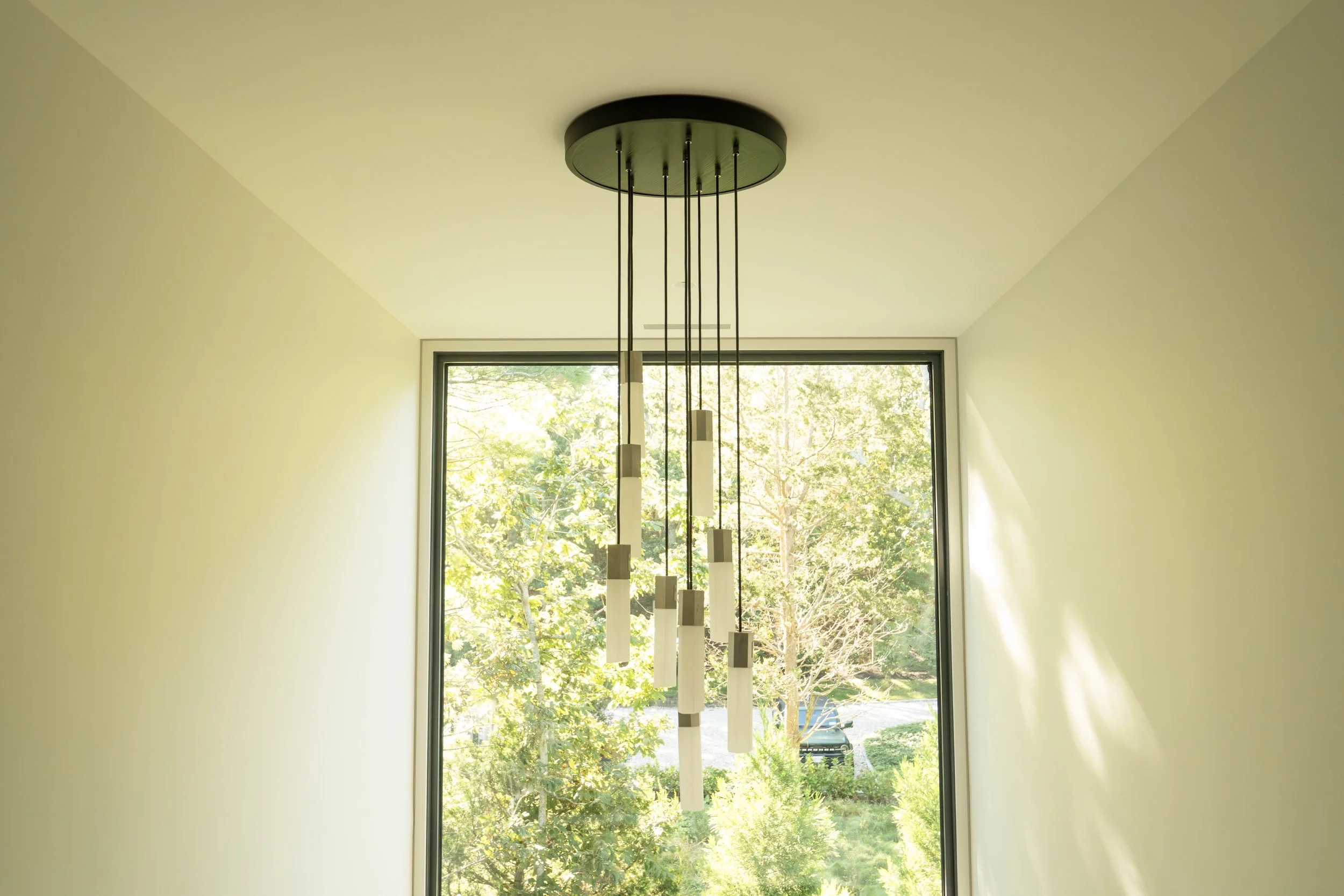 Modern chandelier hanging from ceiling in front of large window with greenery outside.