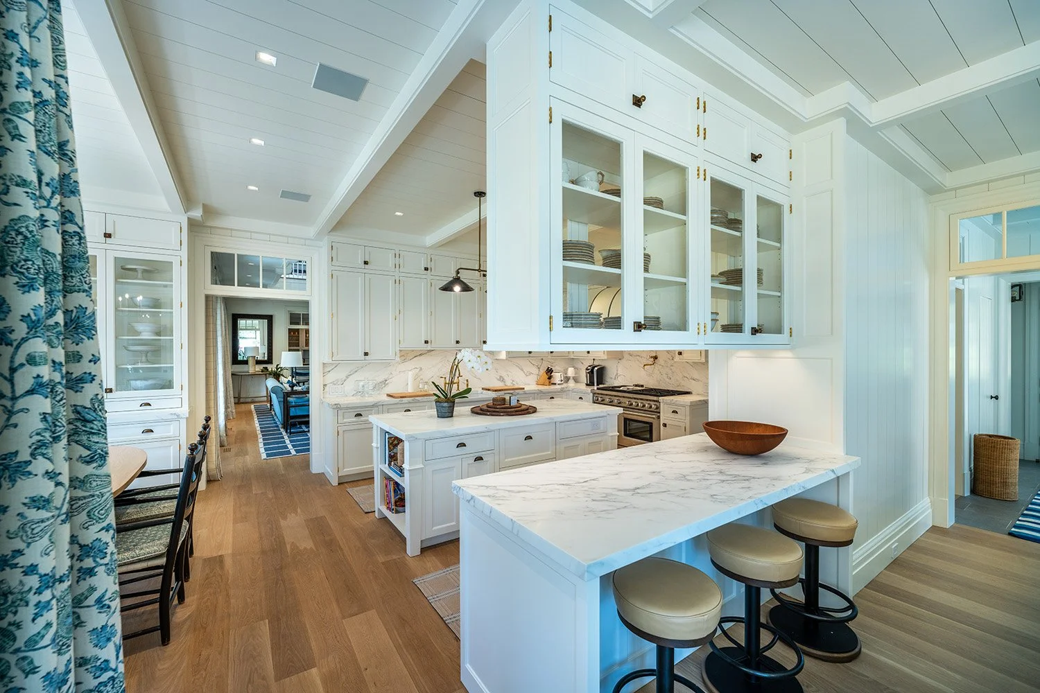 Bright white kitchen with marble countertops, glass-front cabinets, and wooden floors. Visible in the background are a dining area and living room, with blue accents and a doorway leading to a hallway.