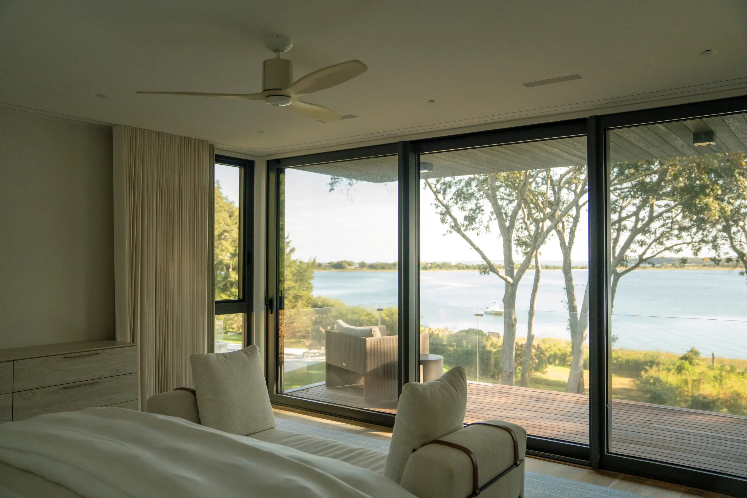 A bedroom with large sliding glass doors overlooking a river, outdoor seating on a deck, trees, and a boat in the water.