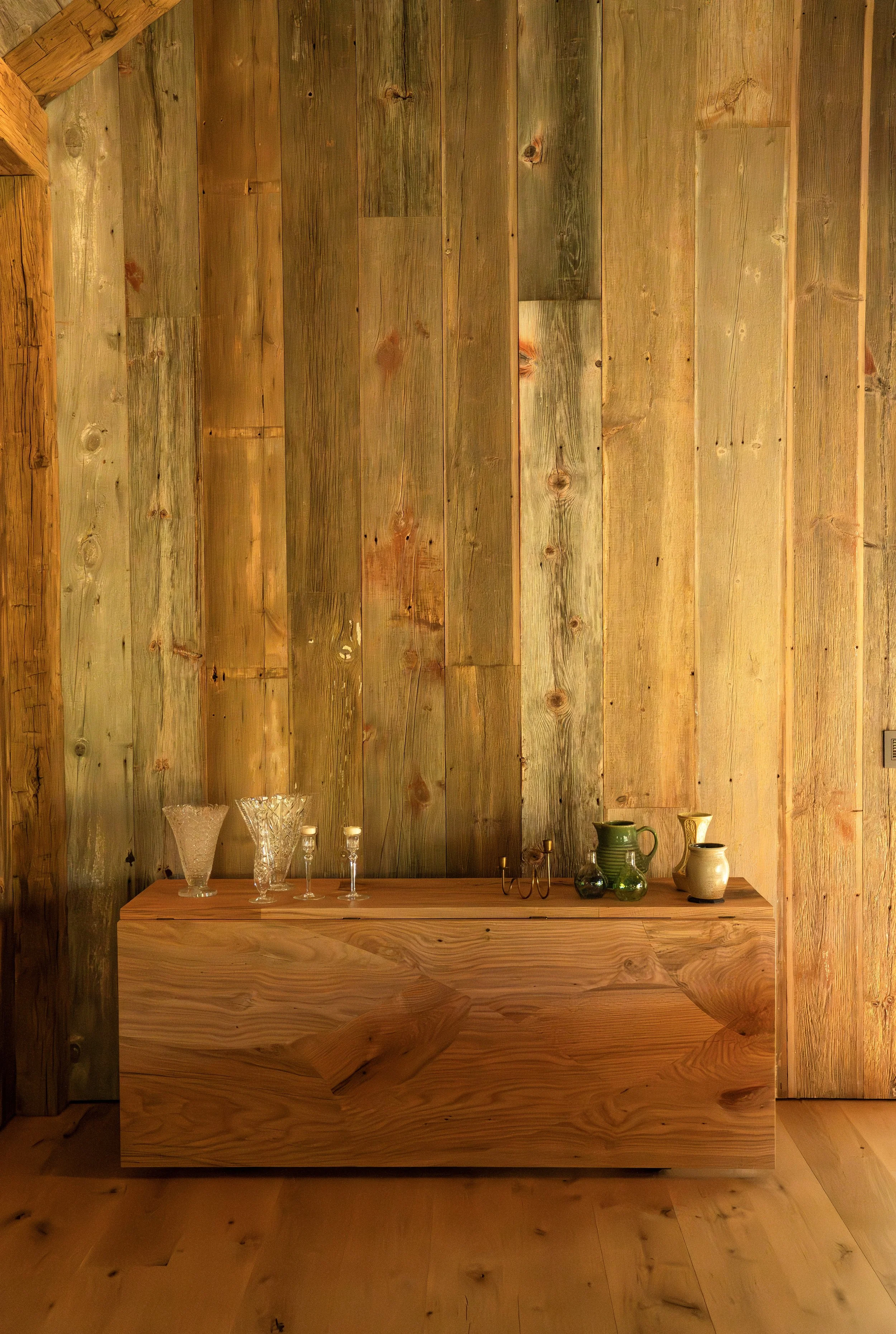 A wooden sideboard against a rustic wooden wall with various glass and ceramic vases and small decorative items on top.