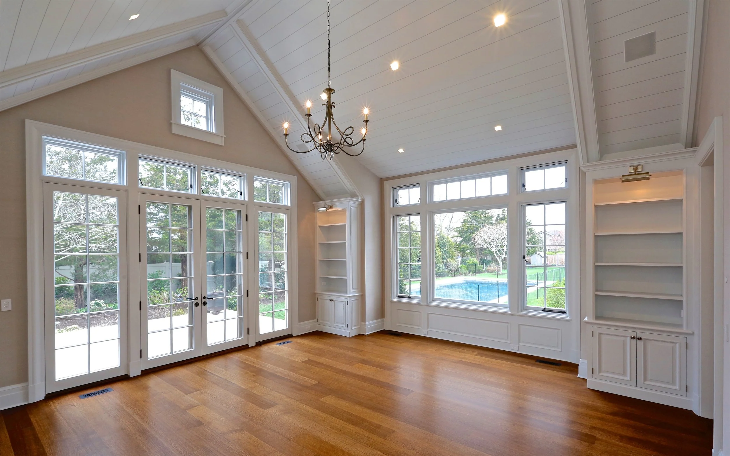 Bright living room with large windows overlooking a backyard with a pool, hardwood flooring, and built-in white bookshelves, featuring a chandelier and recessed lighting.