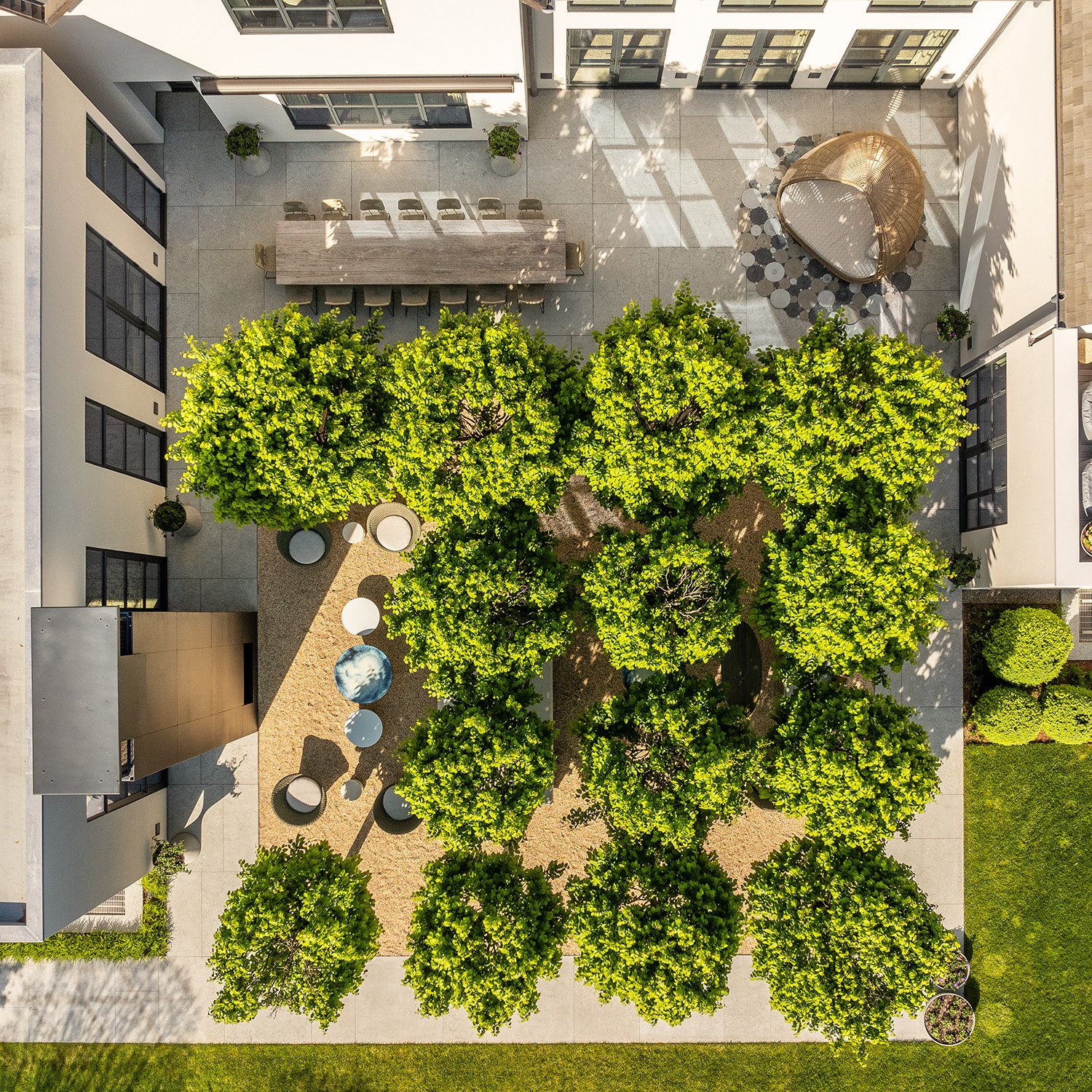 An aerial view of a modern outdoor courtyard with eight green trees centered, surrounded by various furniture and decorative items including a large dining table with chairs, a circular seating area beneath a gold-colored canopy, and several small ta