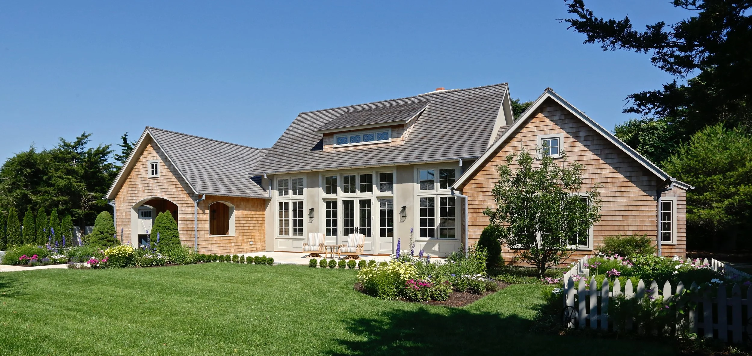 Front view of a large house with wooden shingles, multiple windows, a small porch, and a well-maintained lawn with colorful flower beds and a white picket fence, under a clear blue sky.