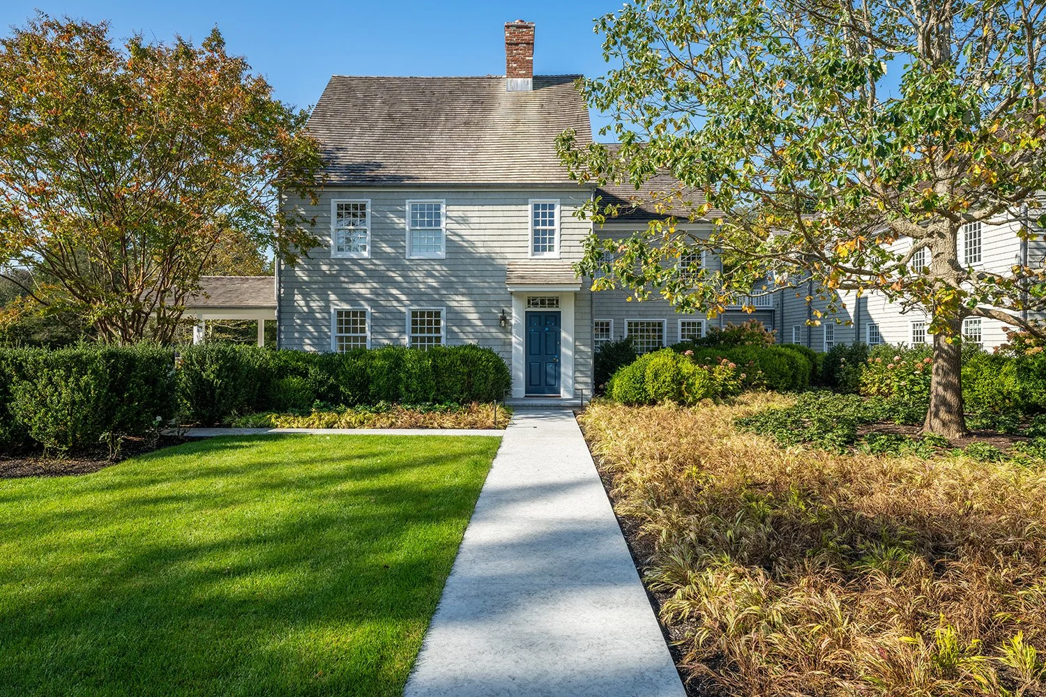 Front view of a gray house with a blue door, surrounded by green bushes, trees, and a manicured lawn, with a concrete walkway leading to the entrance under a clear blue sky.