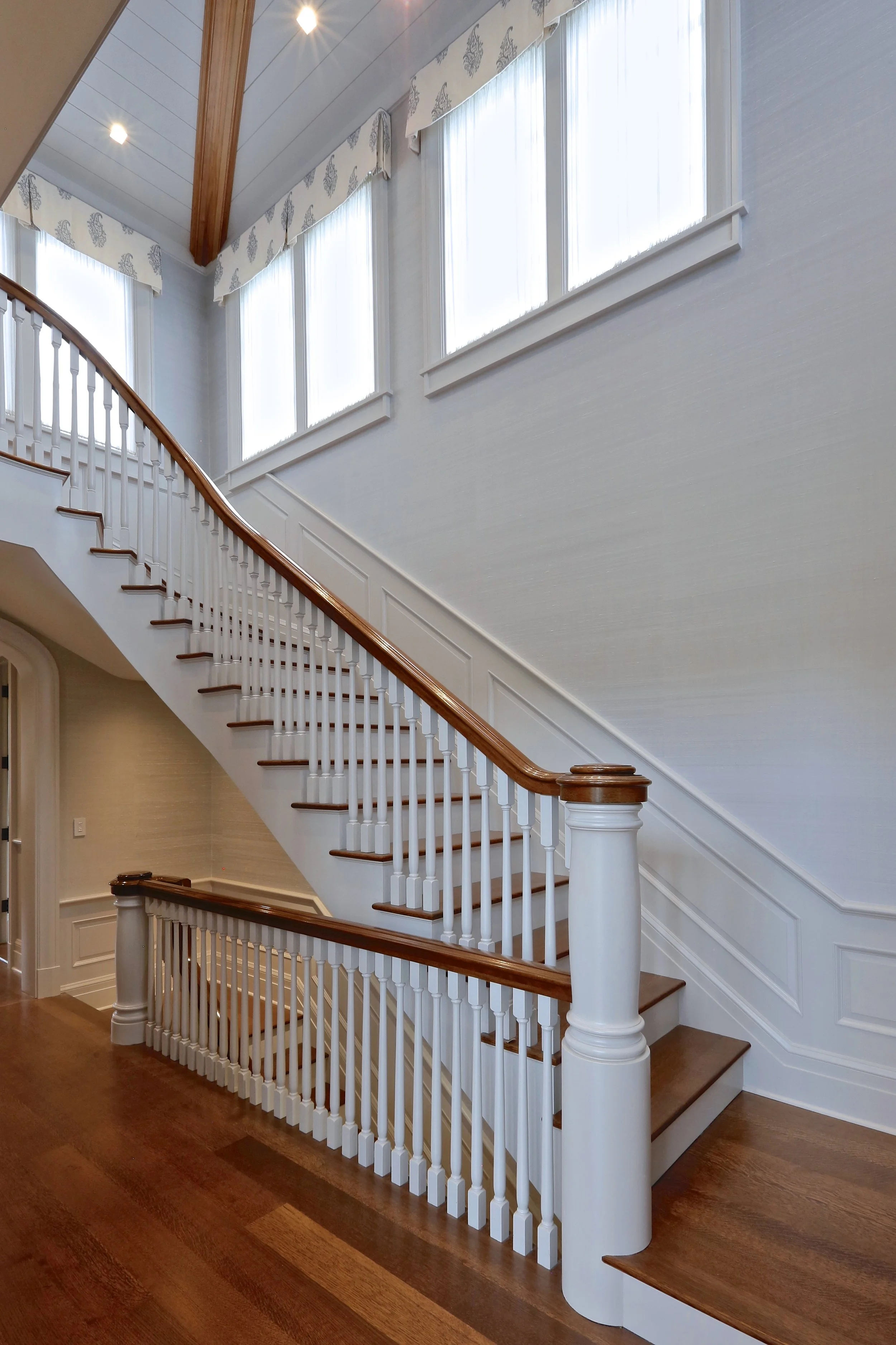 Interior view of a staircase with wooden handrails and white balusters in a house with large windows and wood flooring.