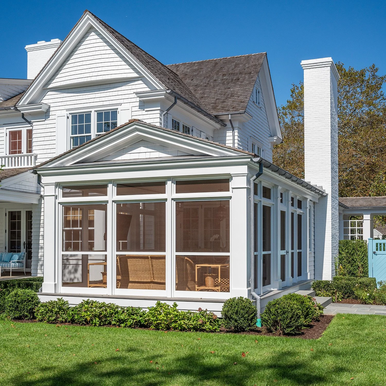 White house with a sunroom enclosed with glass windows, green lawn, shrubs, and clear blue sky.