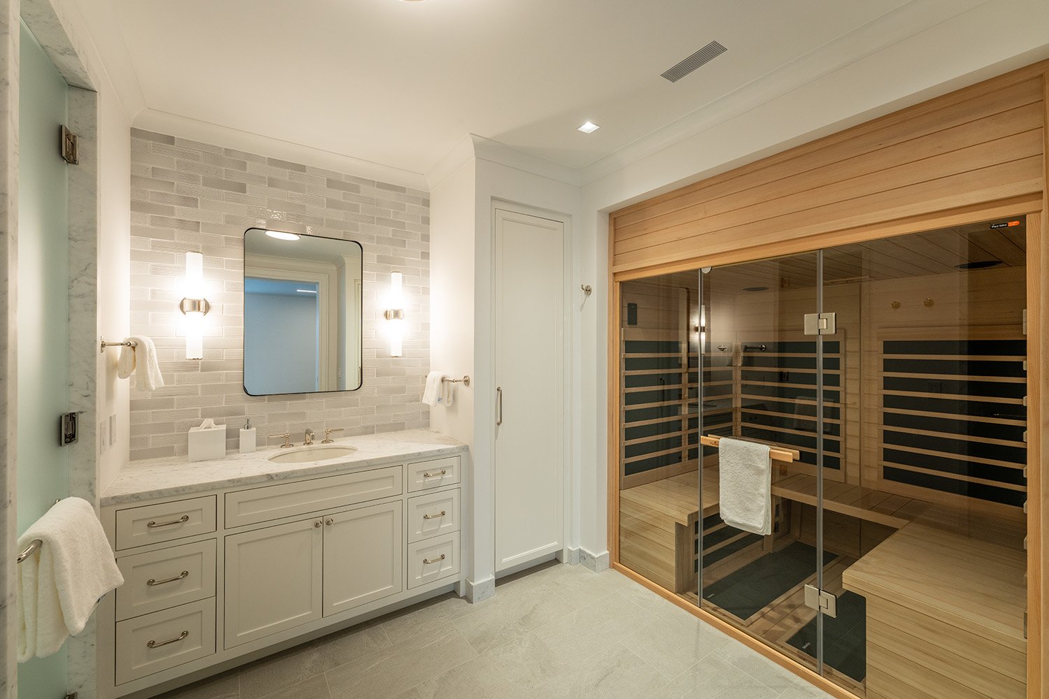 Bathroom with white vanity, mirror, towel racks, and a wooden sauna with glass doors.