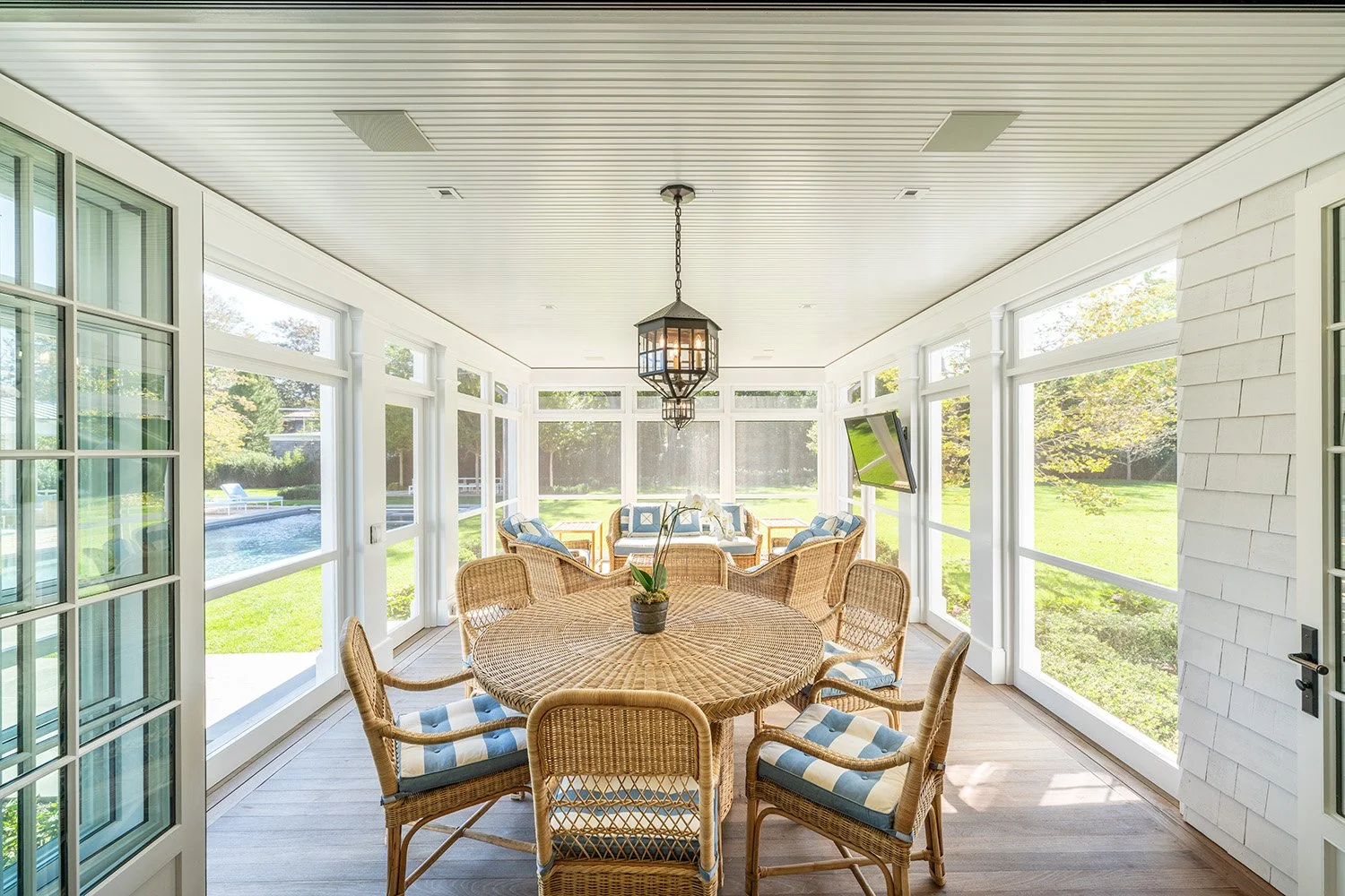 Sunlit screened-in porch with wicker furniture, a round table with chairs, a hanging lantern, wall-mounted TV, and views of a green yard and swimming pool outside.