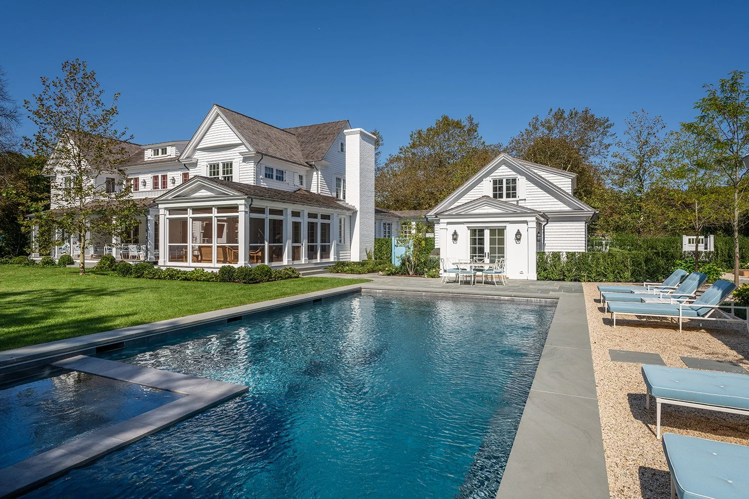 A white residential house with a swimming pool in the backyard, surrounded by green grass, trees, and outdoor lounge chairs.