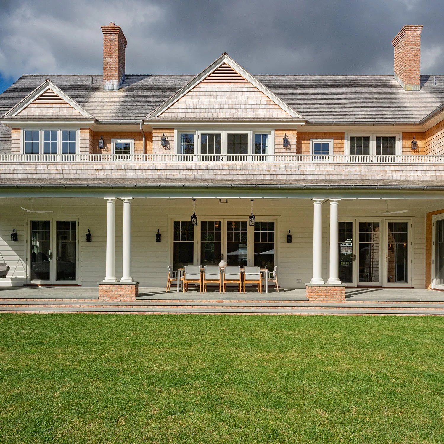 Back view of a large two-story house with a spacious porch and outdoor dining area, green lawn in the foreground, cloudy sky overhead.