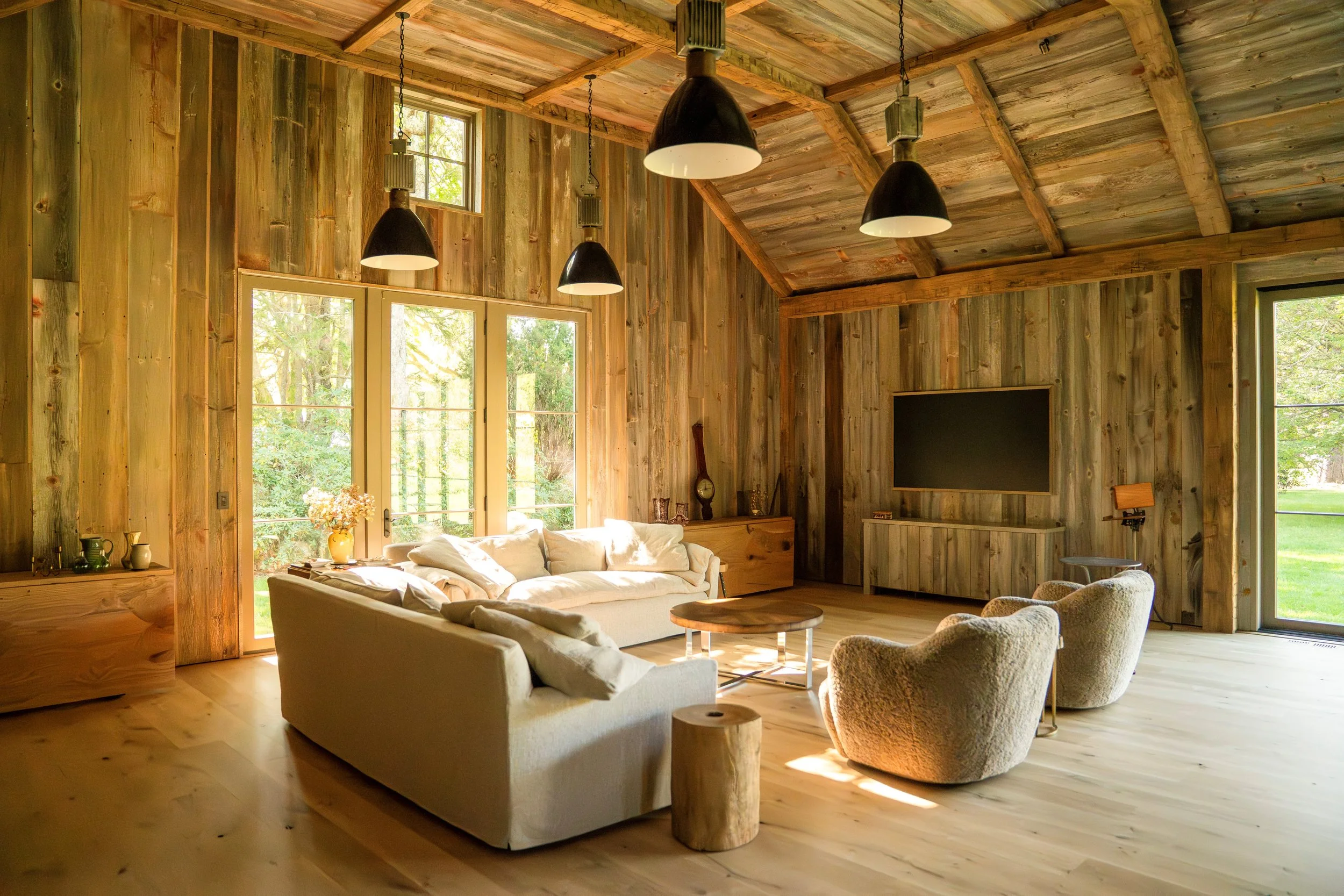 A living room with wooden walls and ceiling, large windows, and modern furniture including a beige sofa, patterned armchairs, a wooden coffee table, and a wall-mounted flat-screen TV. Black pendant lights hang from the ceiling, and sunlight streams t