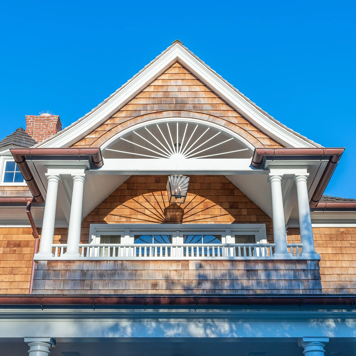 Front view of a house with brown shingles, white columns, a balcony, and a lamp casting a shadow, under a clear blue sky.