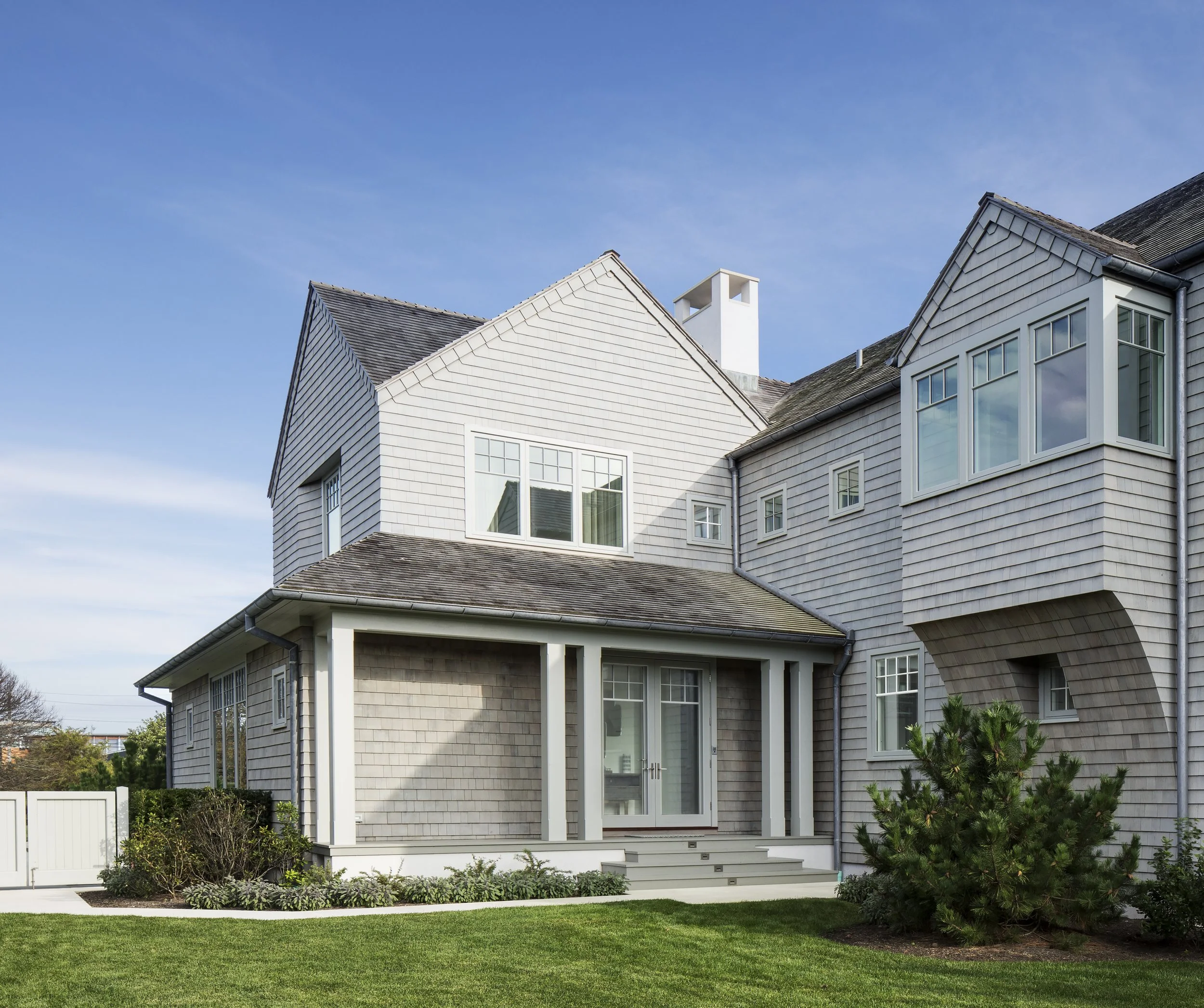 A large multi-story house with gray shingle siding, white trim, and multiple windows, featuring a small covered porch with steps leading to glass doors, surrounded by a well-maintained lawn and shrubbery under a clear blue sky.