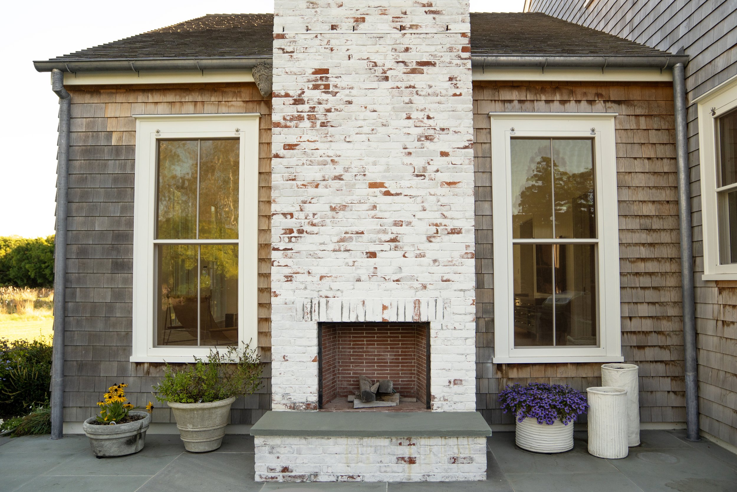 Exterior of a house with a white brick chimney, two windows with white frames, wooden shingles, and potted plants on a stone patio.