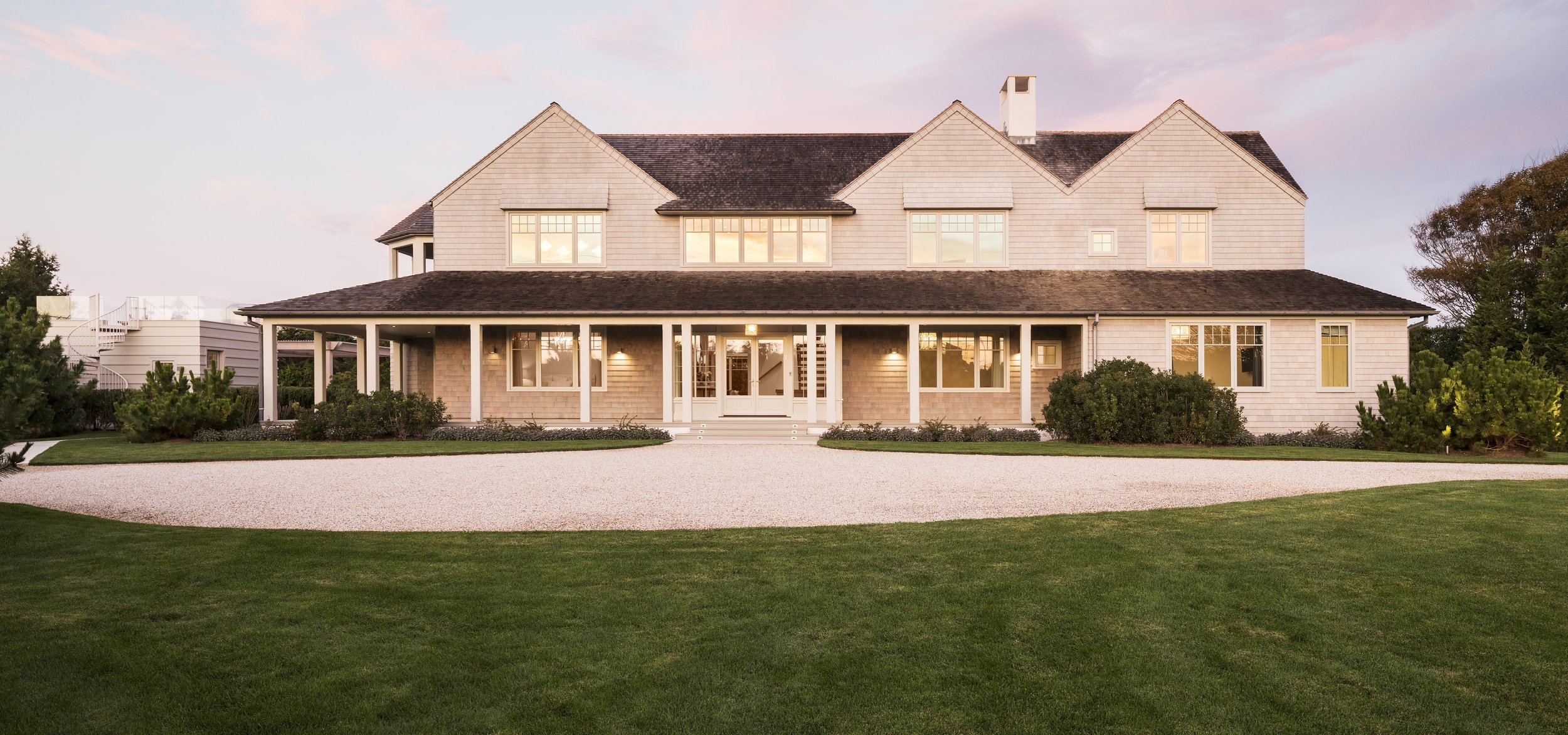 Large two-story house with white exterior, covered porch, multiple large windows, and a dark shingle roof. Surrounded by a well-manicured lawn and bushes, with a gravel pathway in front. Sunset sky in the background.