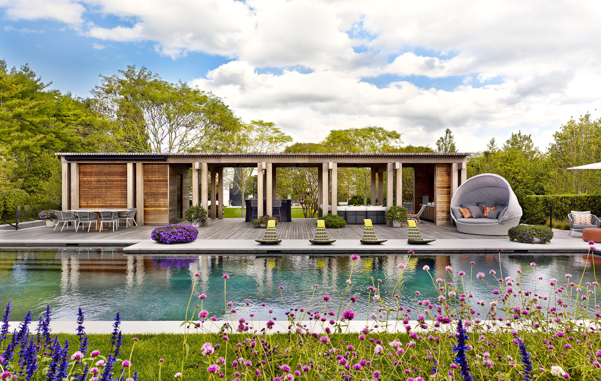 Modern backyard with a pool, outdoor furniture, and a wooden structure, surrounded by colorful flowers and green trees under a partly cloudy sky.
