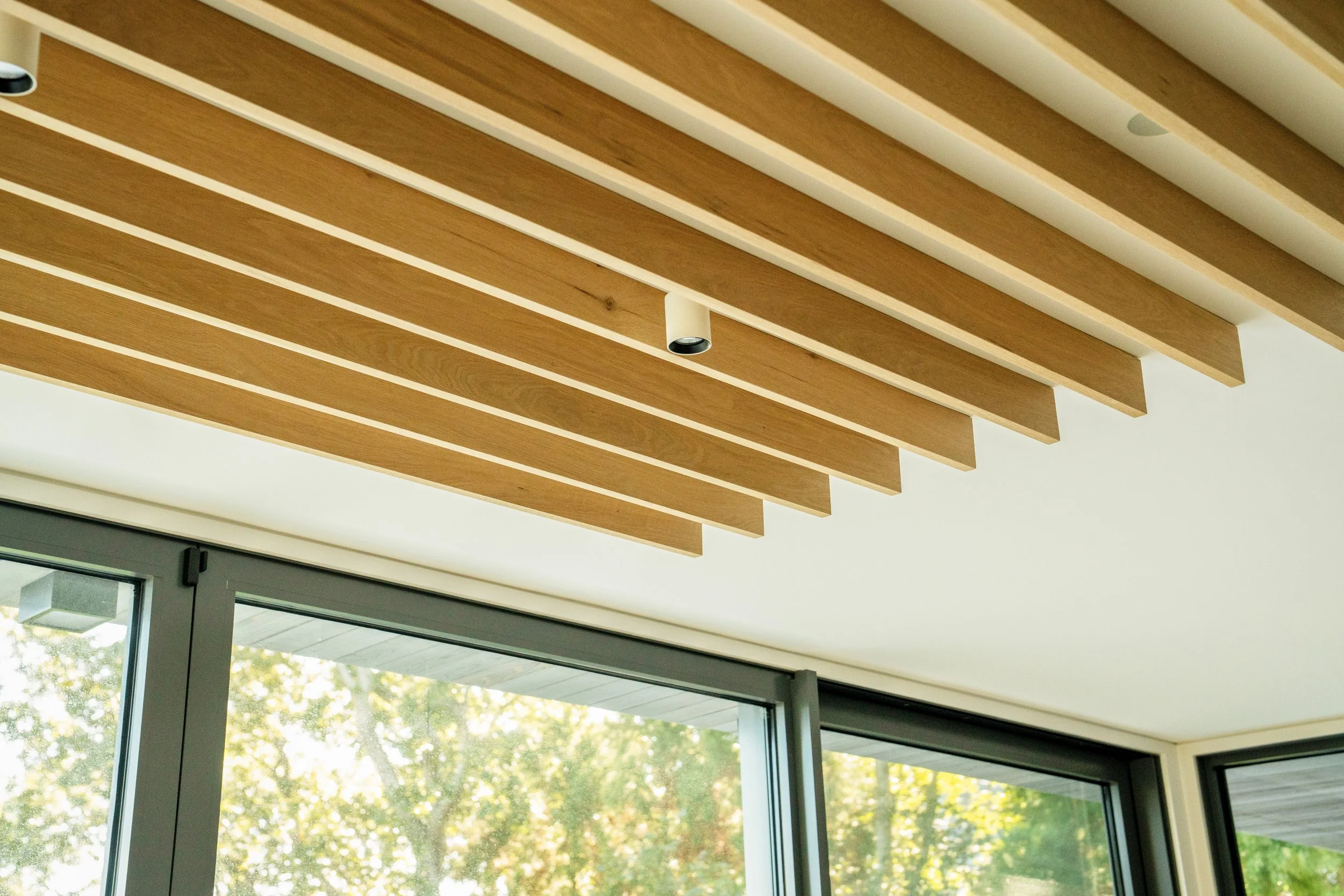 Interior ceiling with wooden slats and embedded lighting, next to large glass windows showing trees outside.