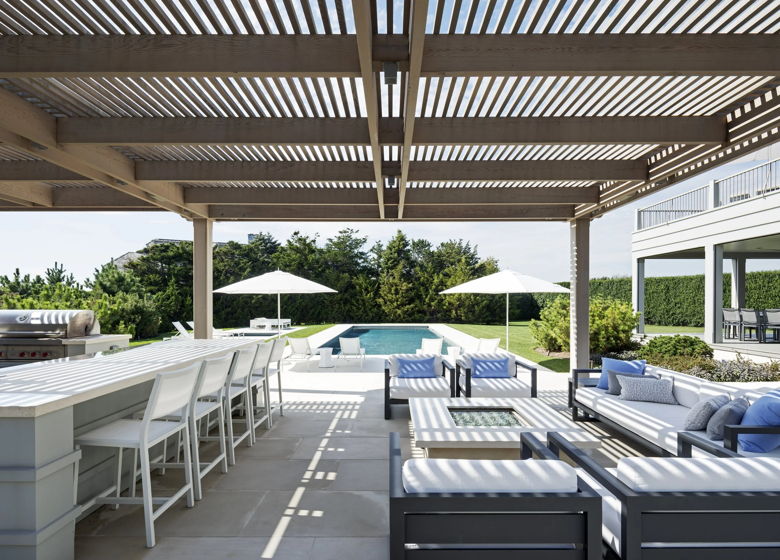 Outdoor patio area with seating and a swimming pool, shaded by a wooden pergola with slatted roof, on a sunny day.