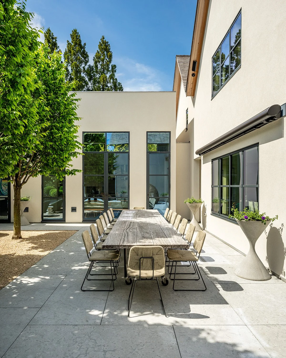 Outdoor patio area with a long wooden dining table and 12 beige chairs, surrounded by modern white buildings with large windows, potted plants, and a leafy tree, on a sunny day.