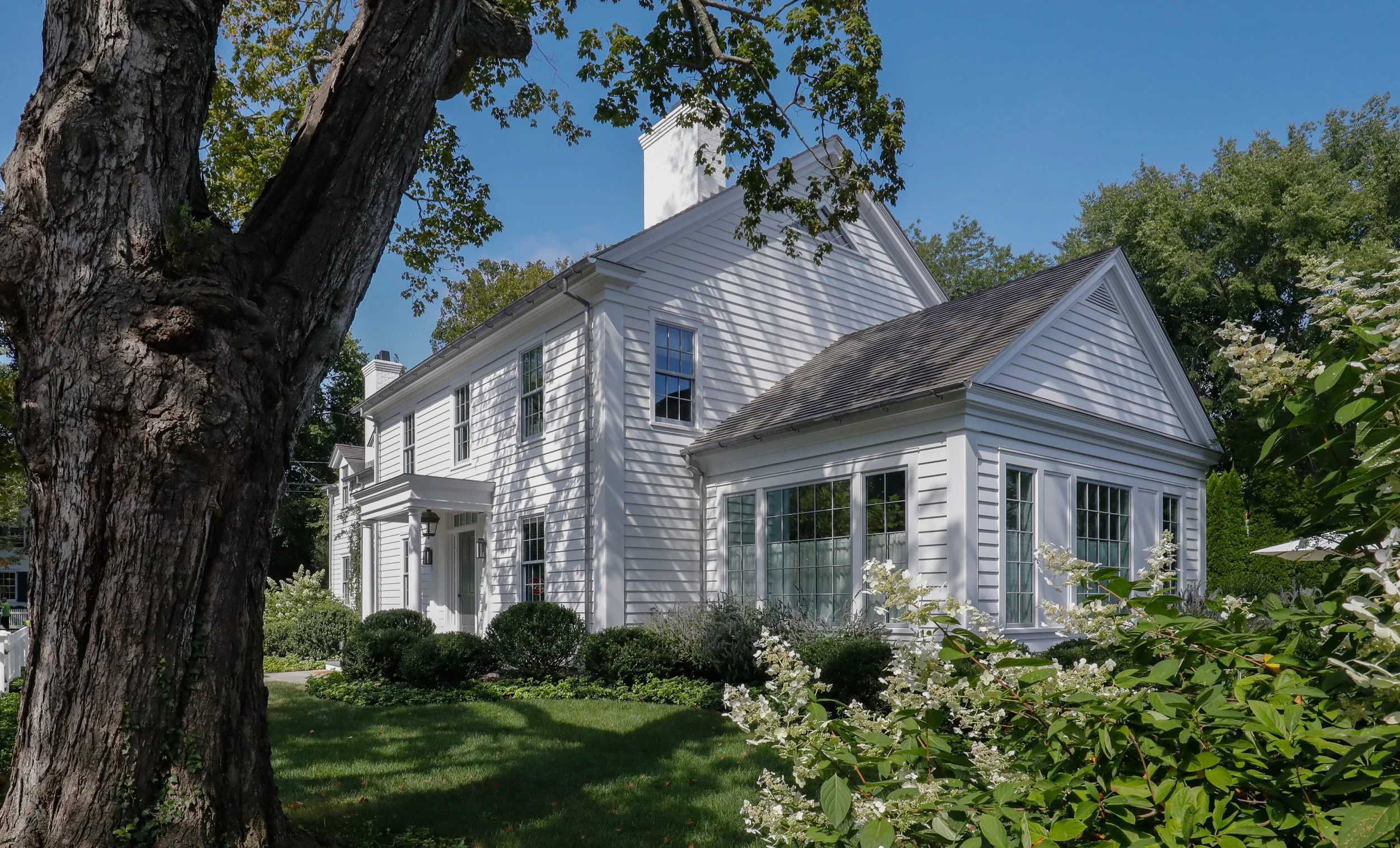 A white, two-story house with multiple windows, surrounded by green bushes and trees, on a bright, sunny day. Construction in the Hamptons