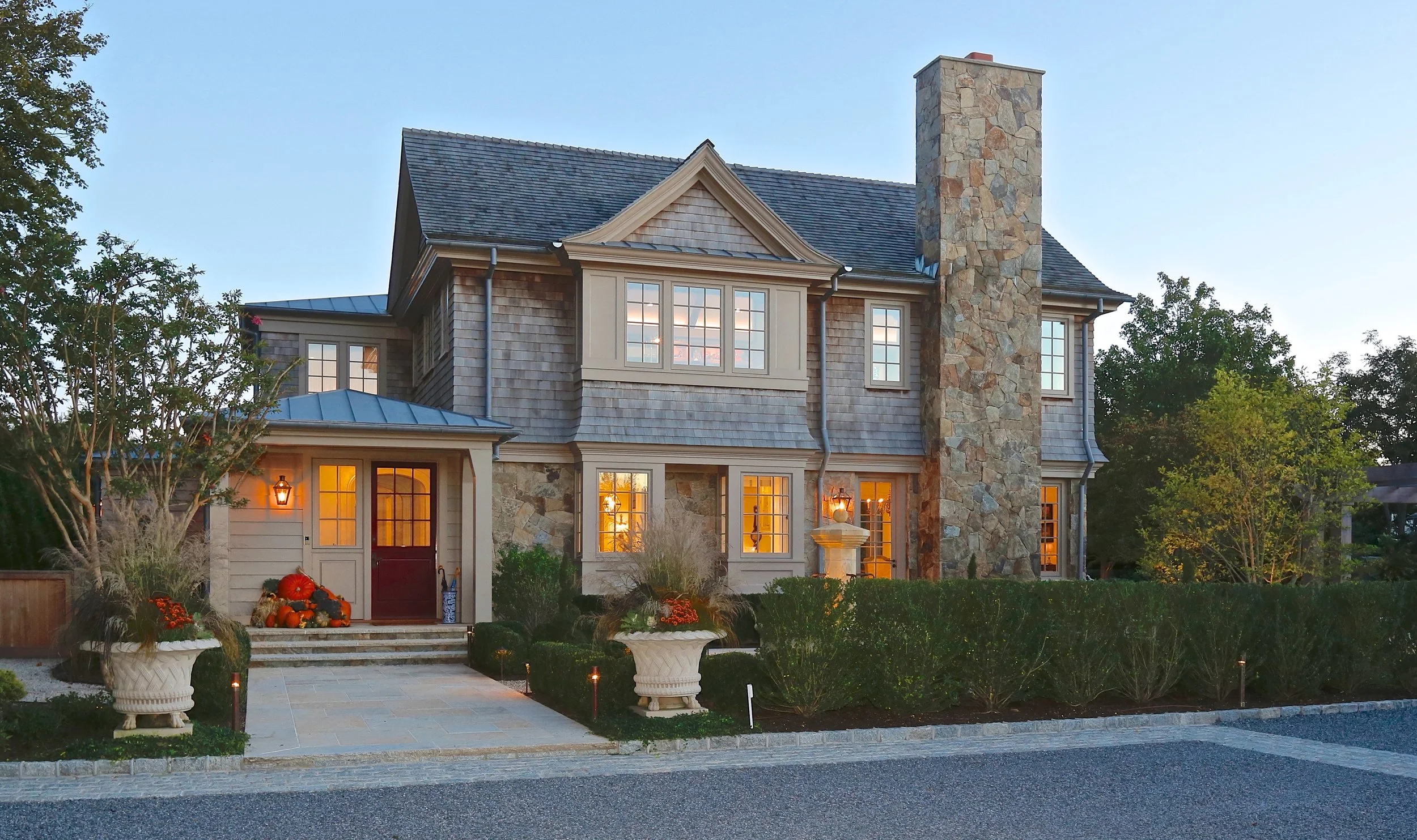 A large two-story house with a stone chimney, lit windows, a stone and wood exterior, and front steps decorated with pumpkins and fall plants during dusk.
