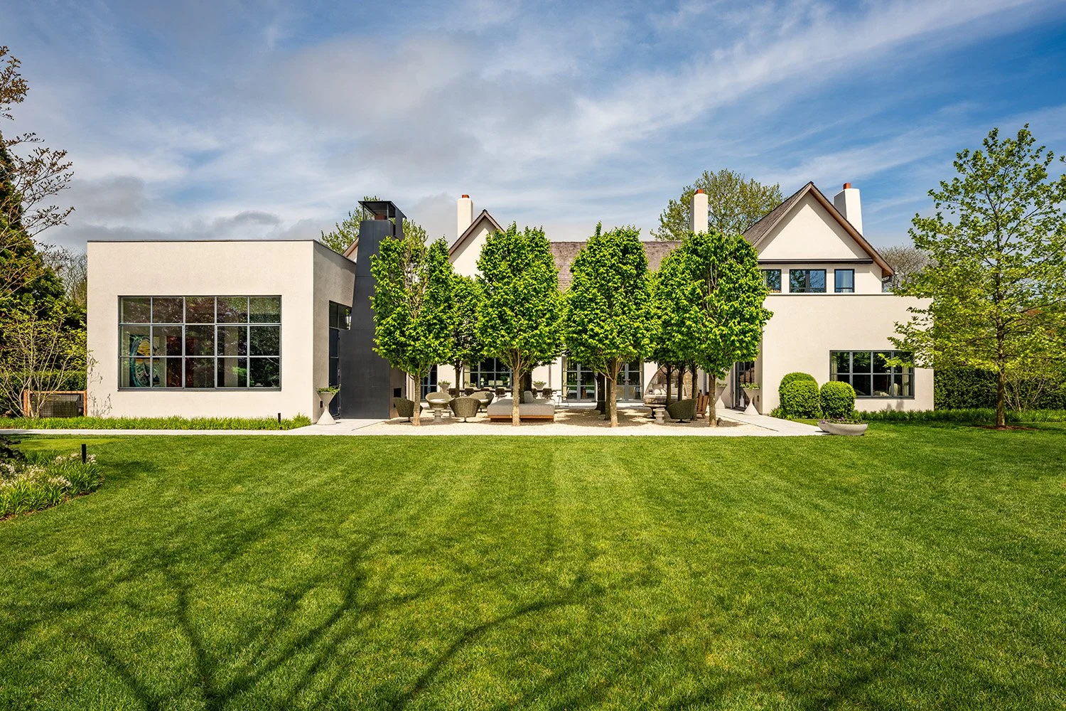 Modern house with large windows, surrounded by a green lawn and trees, with a blue sky above.