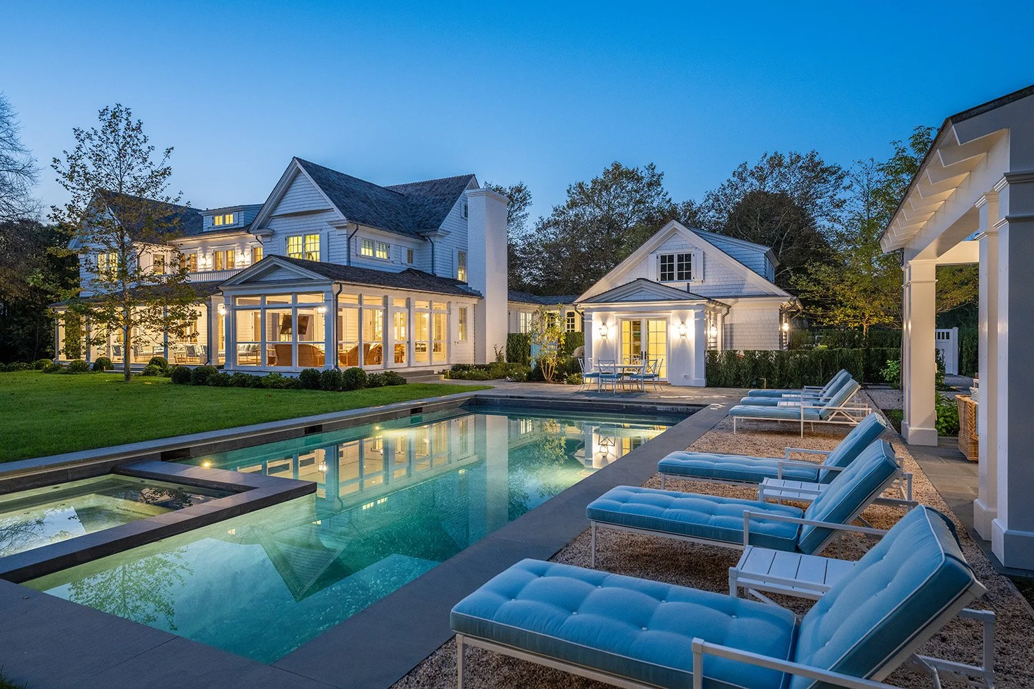 A luxurious backyard with a swimming pool, sun loungers, and a well-lit house with large windows and a patio at dusk.